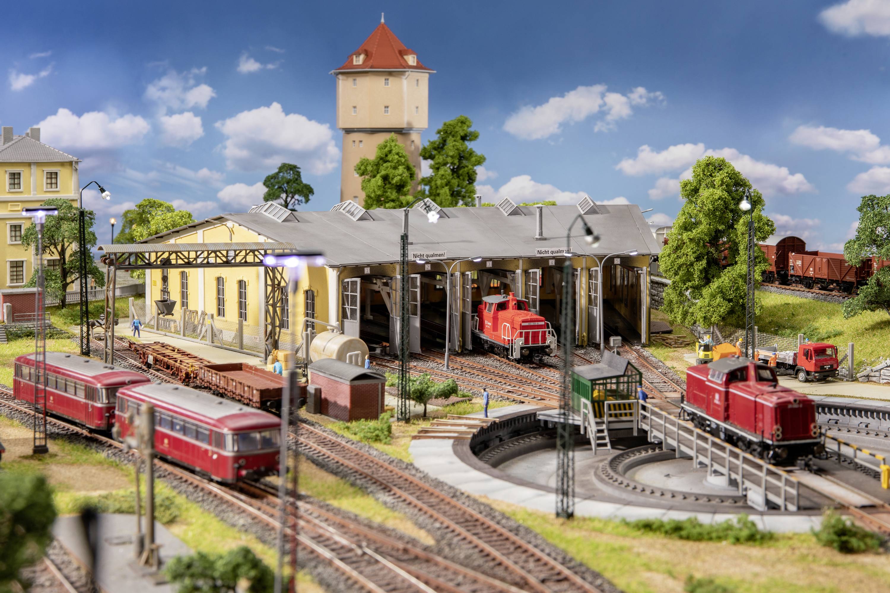 Model kolejiště s lokomotivní remízou, několika kolejemi, dvěma červenými vlaky, vodní věží v pozadí a zelenou krajinnou dekorací.