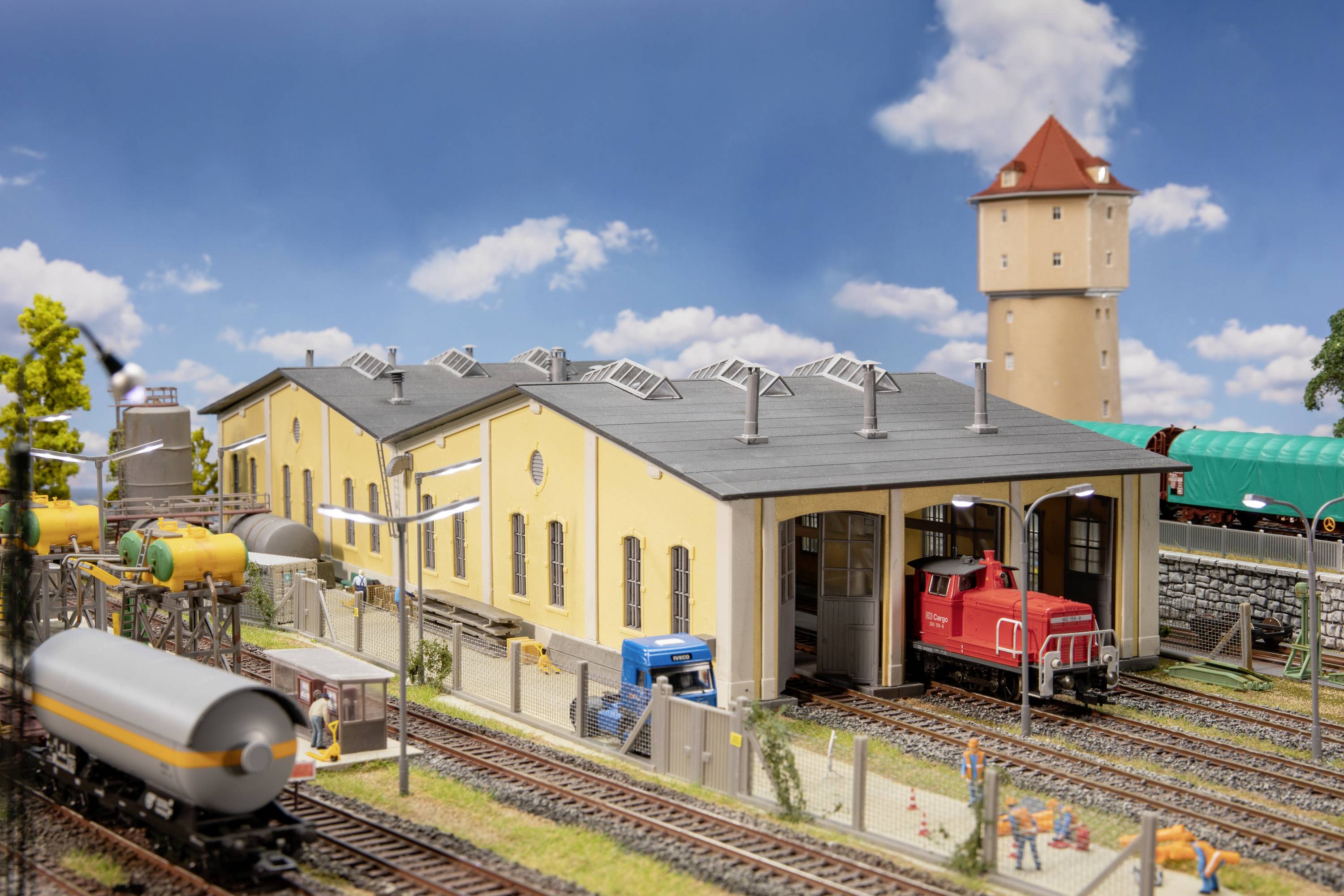 Model vláčkové kolejiště znázorňuje průmyslovou oblast s žlutou tovární budovou, červeným vlakem, několika kolejemi a vodárenskou věží.