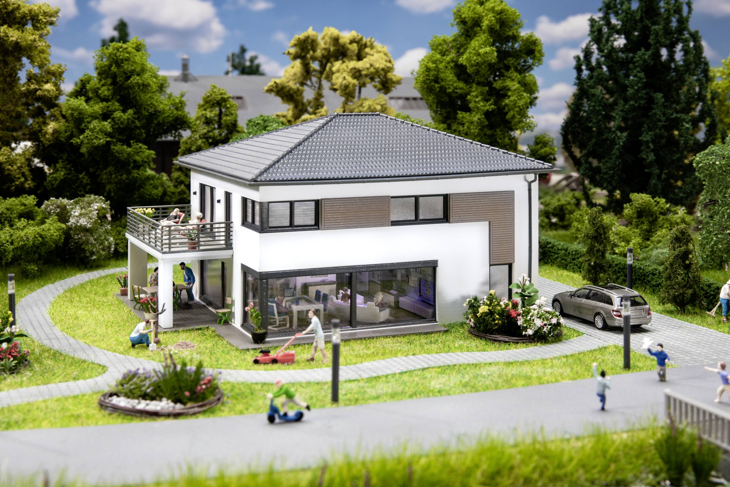 Moderní model rodinného domu se zahradou, obklopený miniaturními stromy a figurkami na cestičce.