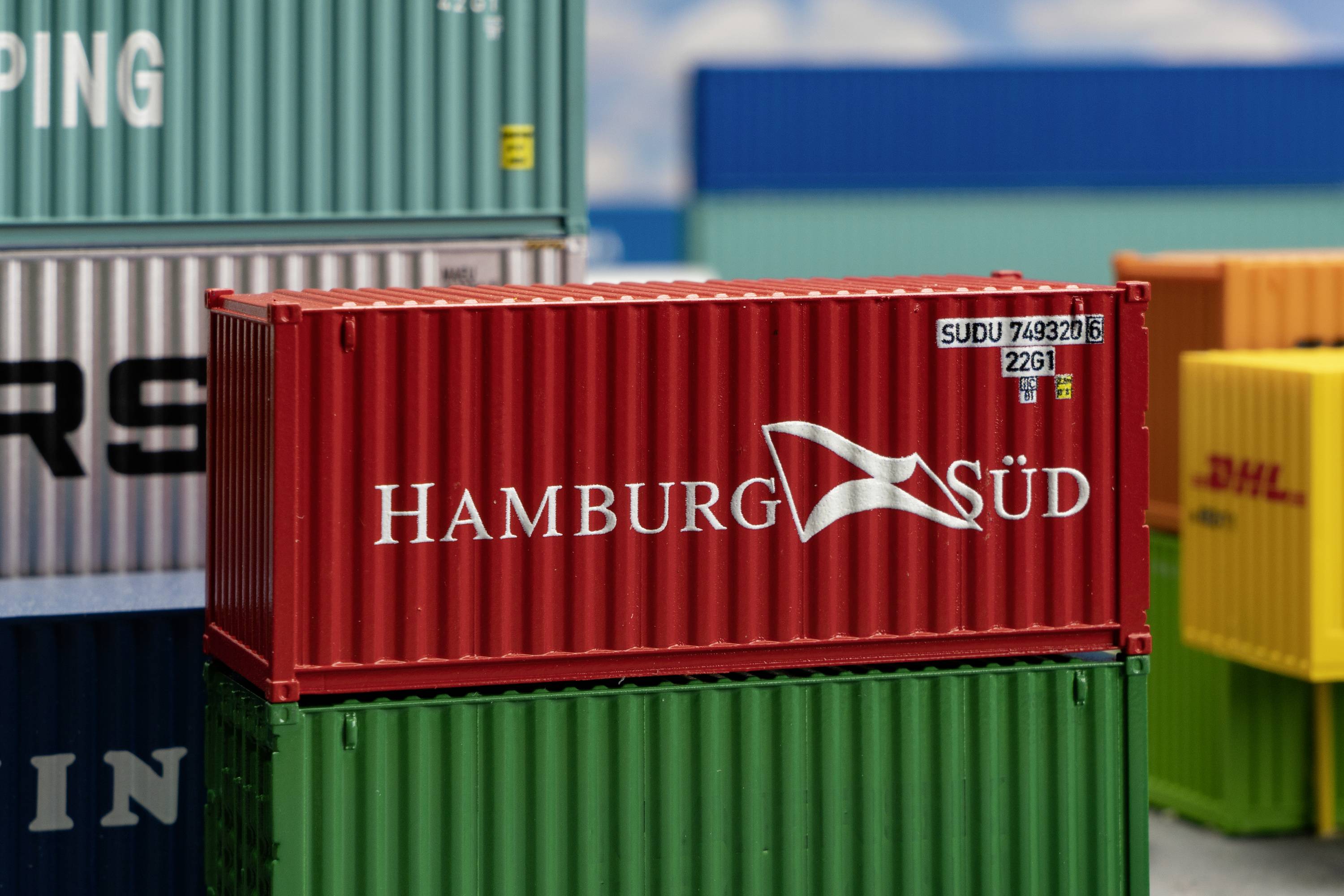 Červený nákladní kontejner s logem společnosti 'Hamburg Süd' umístěný na hromadě dalších kontejnerů v různých barvách, z části viditelných.