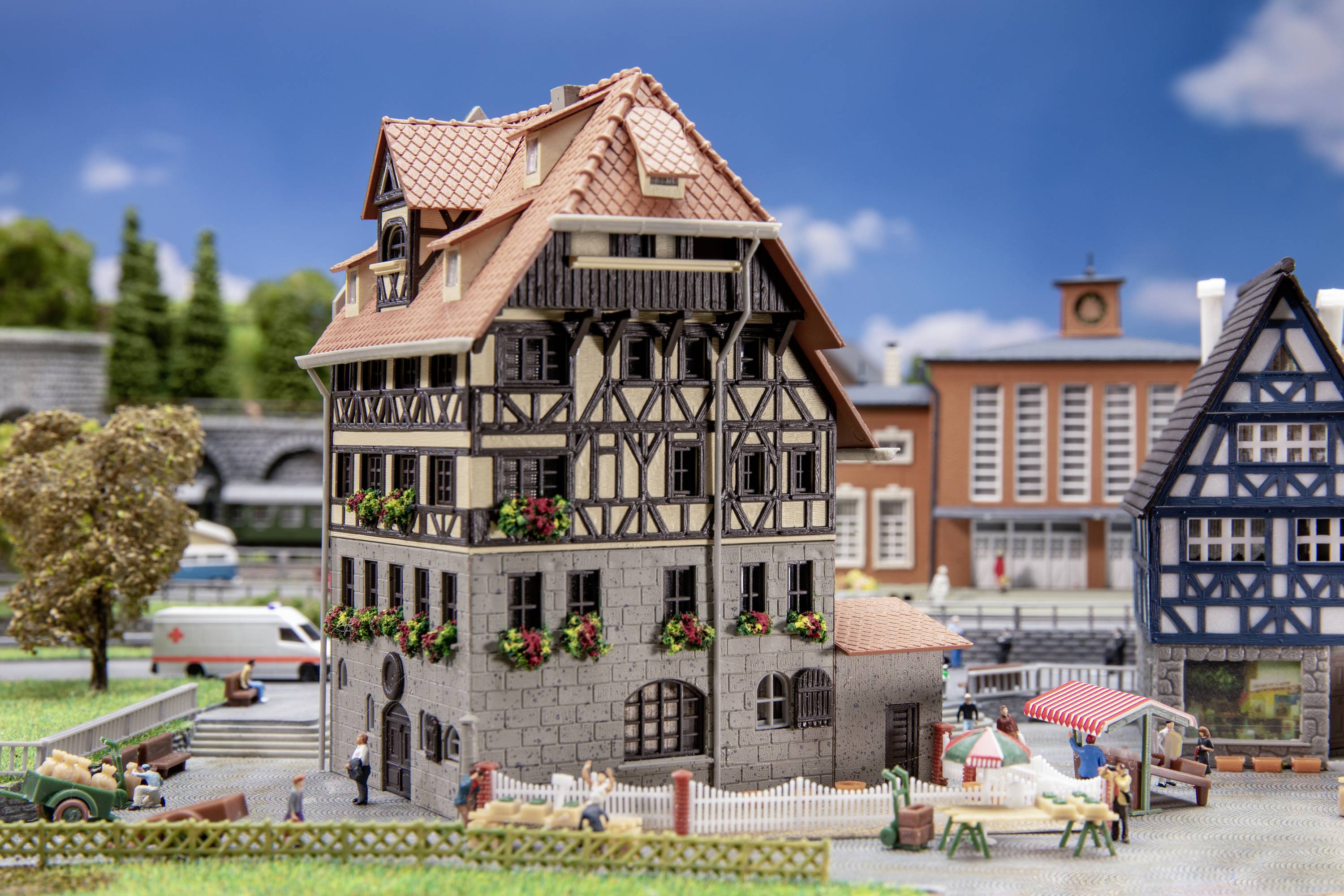 Miniaturní model evropského města zobrazuje hrázděný dům s truhlíky na květiny, obklopený tržními stánky a lidmi.