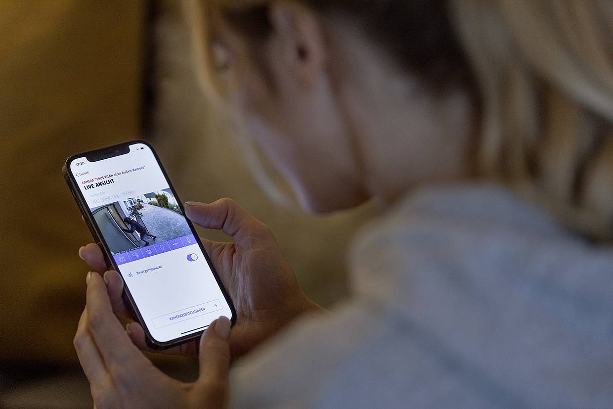 Jedna osoba sleduje smartphone zobrazující živý přenos z bezpečnostní kamery. Kamera snímá vstup do domu.