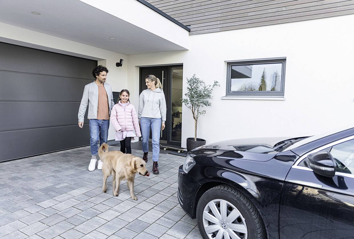 Rodina se psem se usmívá, když kráčí po dlážděné cestě před moderním domem. V popředí je zaparkované černé auto.