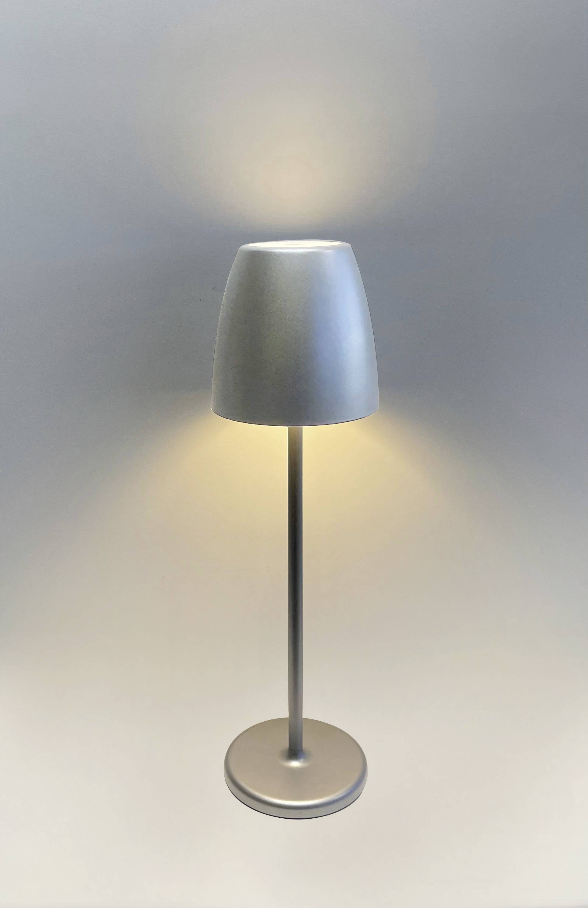 Moderní stříbrná stolní lampa s jednoduchým válcovitým stínidlem, která jemně osvětluje strop a podlahu.