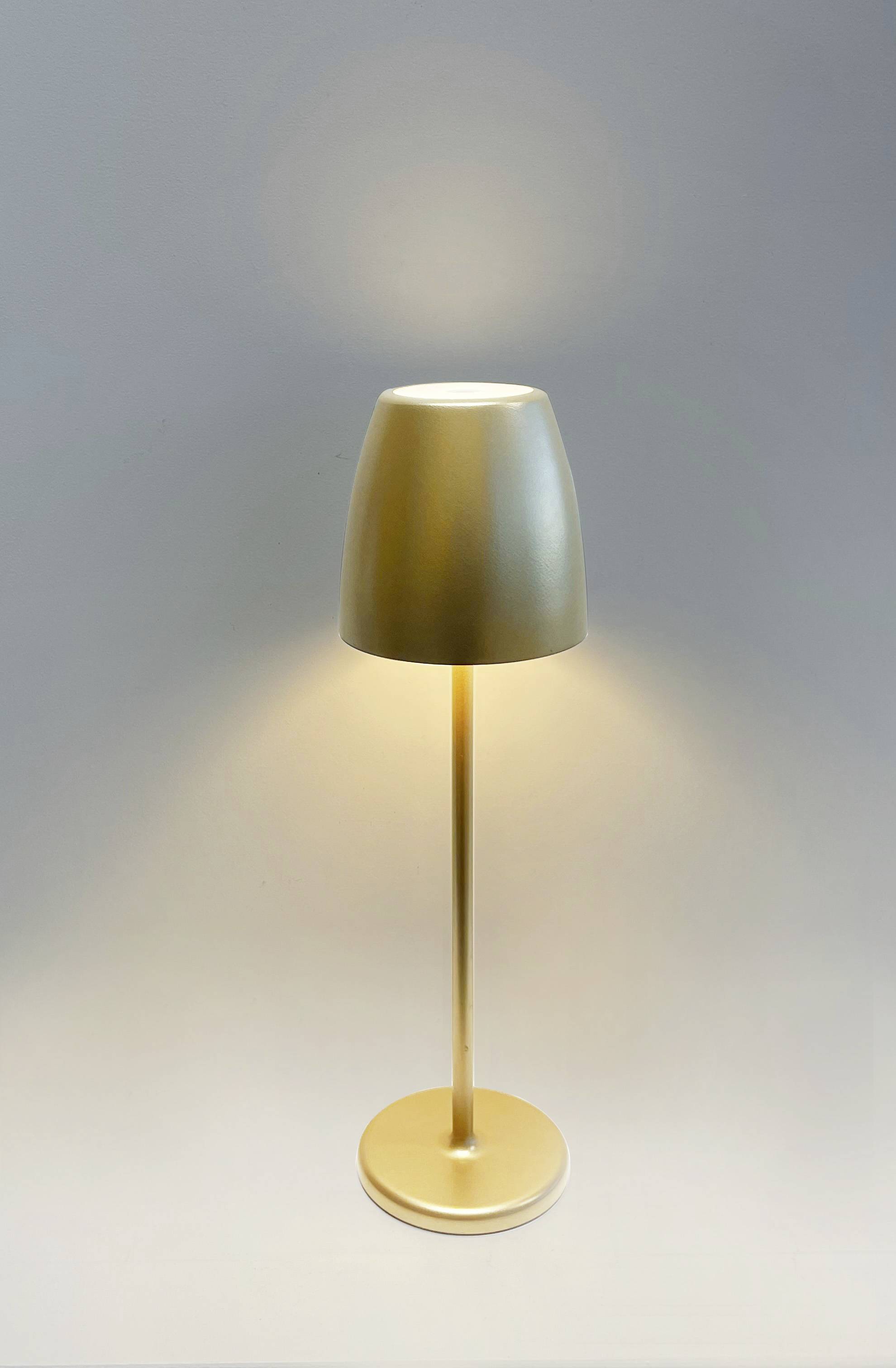 Zlatá stolní lampa s válcovou základnou a kuželovitým stínítkem jemně osvětluje neutrální prostor.