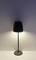 Černá stojací lampa svítí na bílé stěně. Světlo vyzařuje nahoru i dolů a zajišťuje jemné osvětlení.