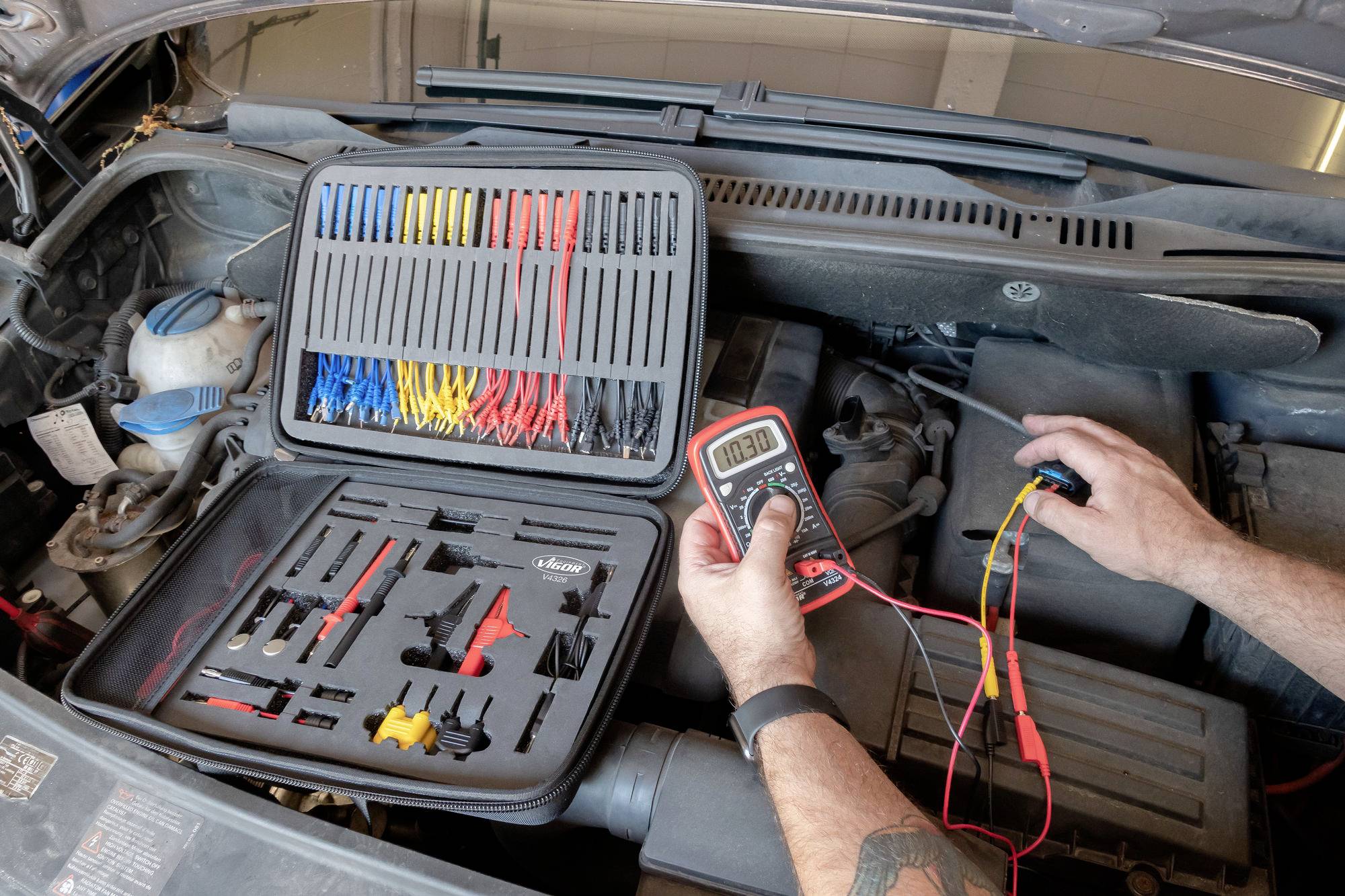 Mechanik používá multimetr ke kontrole elektrické soustavy automobilu. V popředí je viditelná otevřená sada nástrojů.
