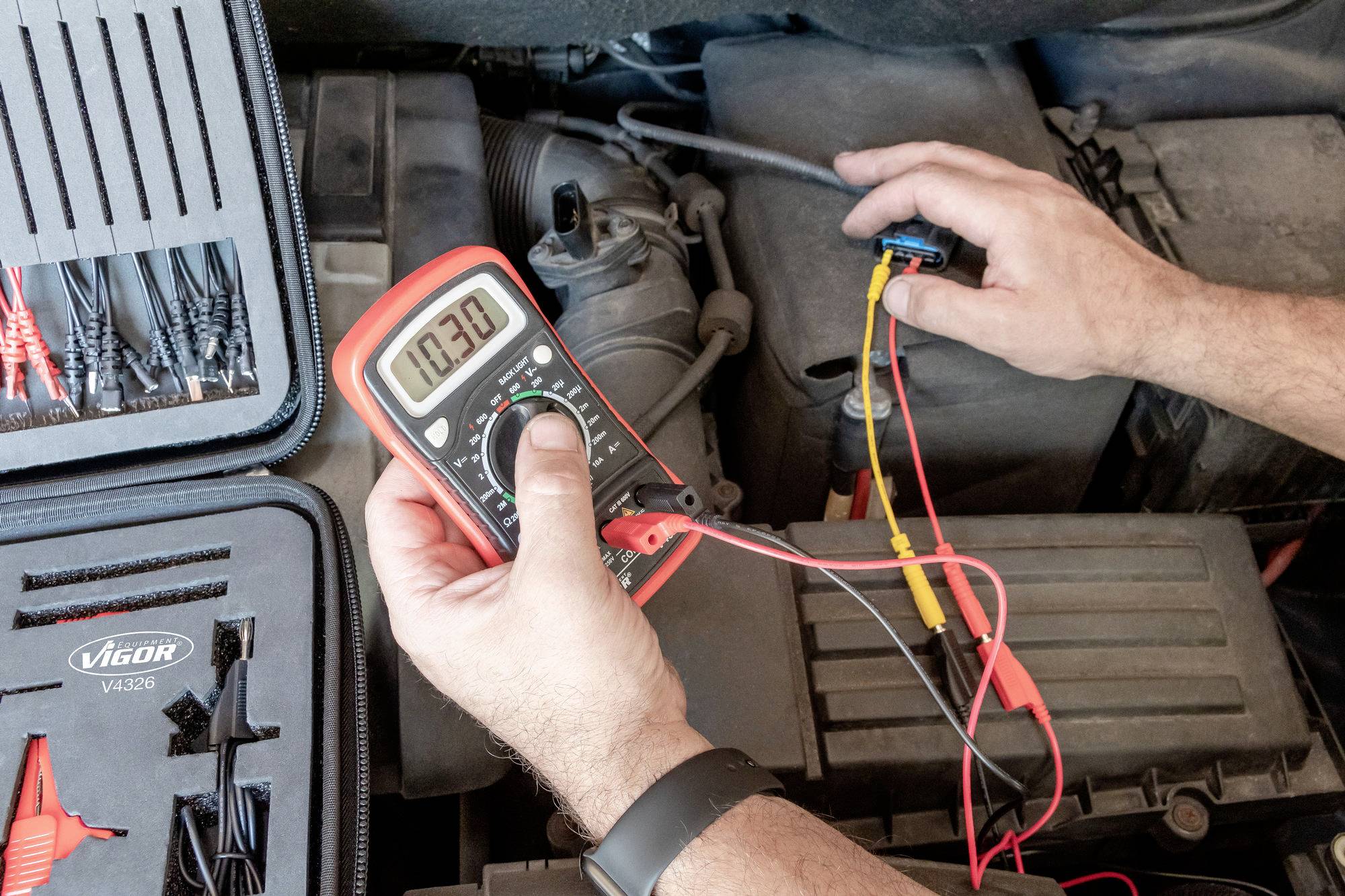 Elektrikář měří napětí autobaterie multimetrem. Vedle motorového prostoru leží přepravka s nástroji a kabely.