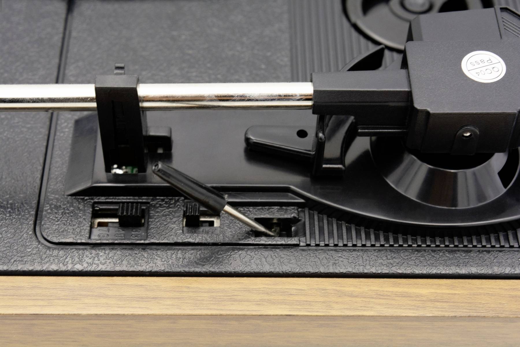 Rameno gramofonu a jehla nad vinylovou deskou, připravené ke spuštění. Mechanické detaily a dřevěný rám jsou viditelné.