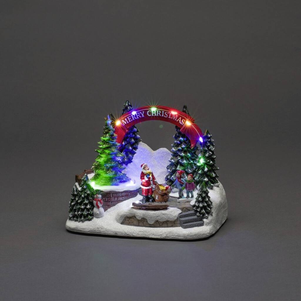 Vánoční dioráma s osvětlenými vánočními stromky, Santa Clausem se sáněmi a zářícím obloukem s nápisem 'Merry Christmas'.