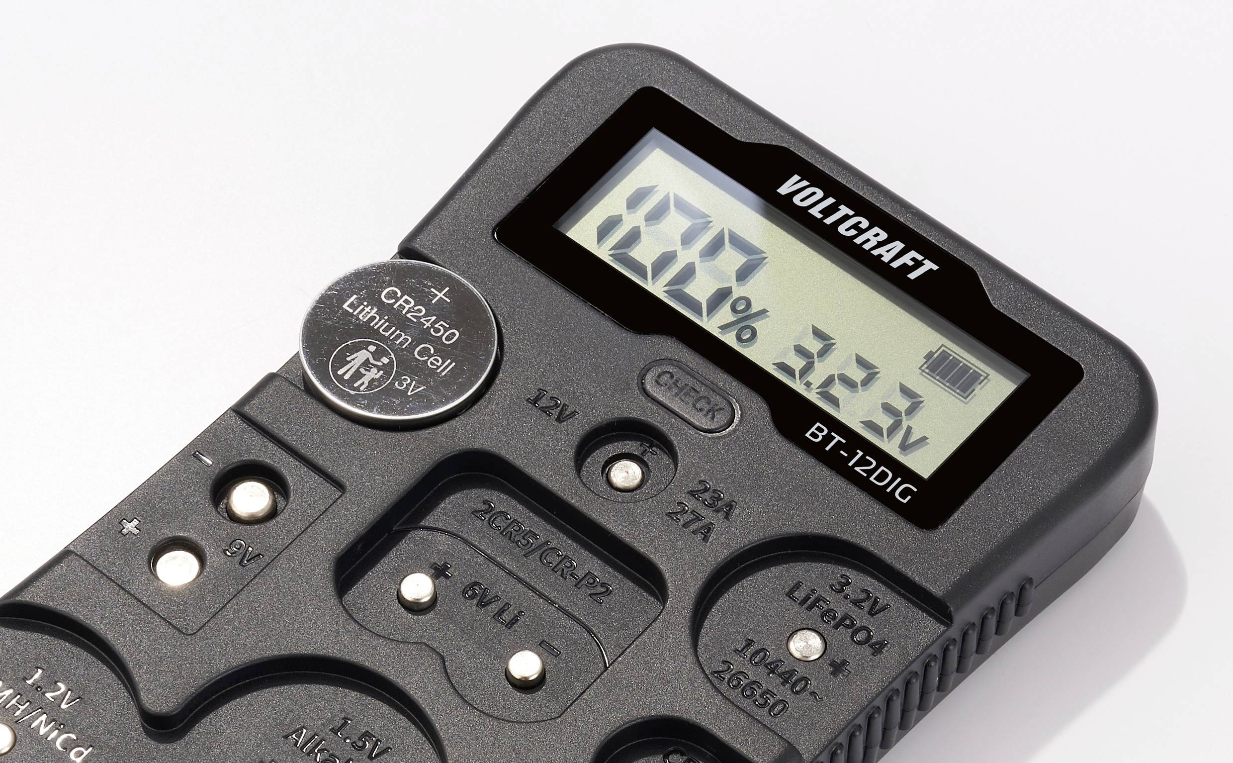 Digitální tester baterií zobrazuje na displeji '100%'. Obsahuje různé sloty pro baterie různých typů.