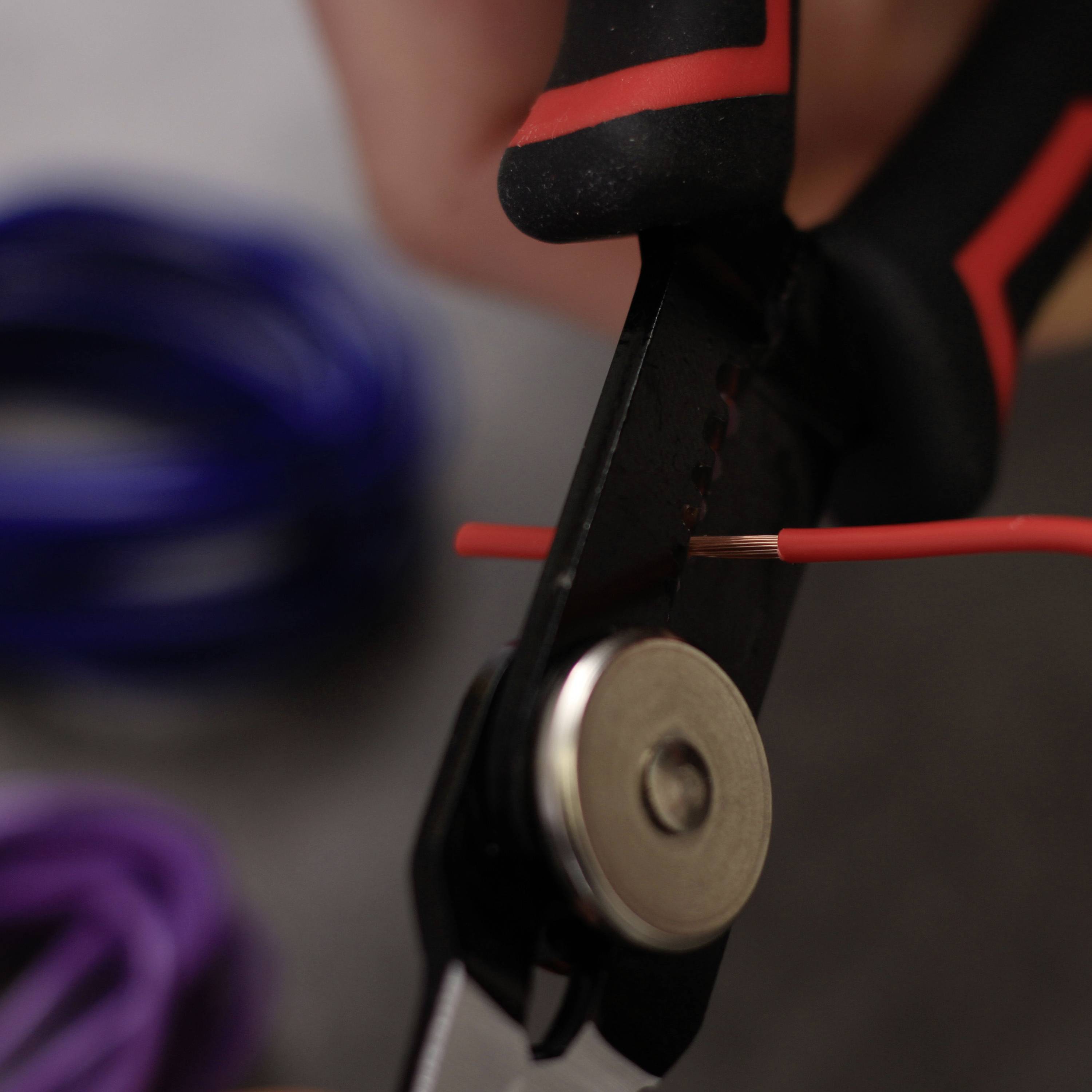 Osoba stříhá tenký černý kabel malými kleštěmi. Kleště mají červený a černý úchop.