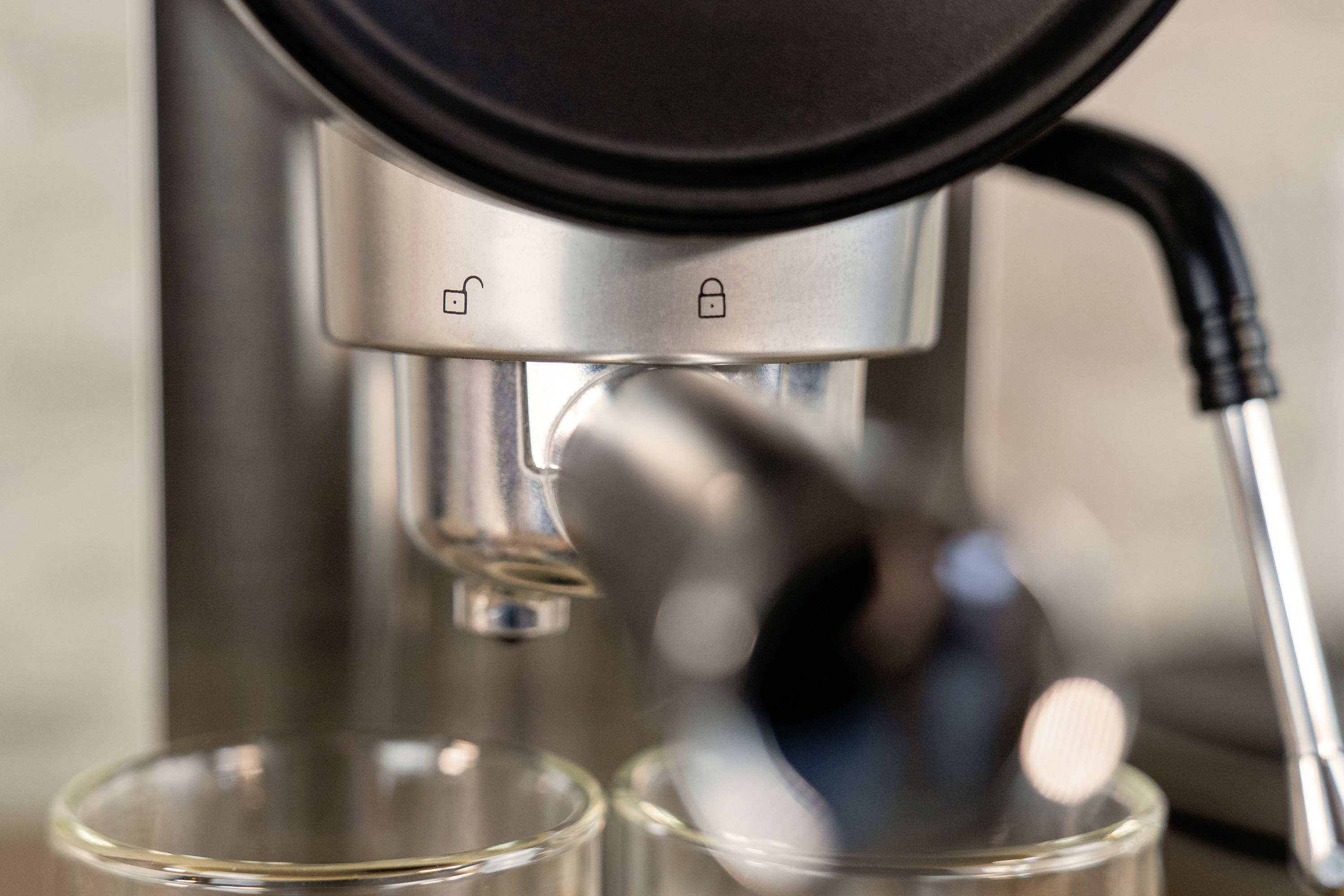 Detailní záběr espresso stroje, ze kterého vytéká káva do dvou sklenic. Symboly indikují, zda je stroj uzamčen.