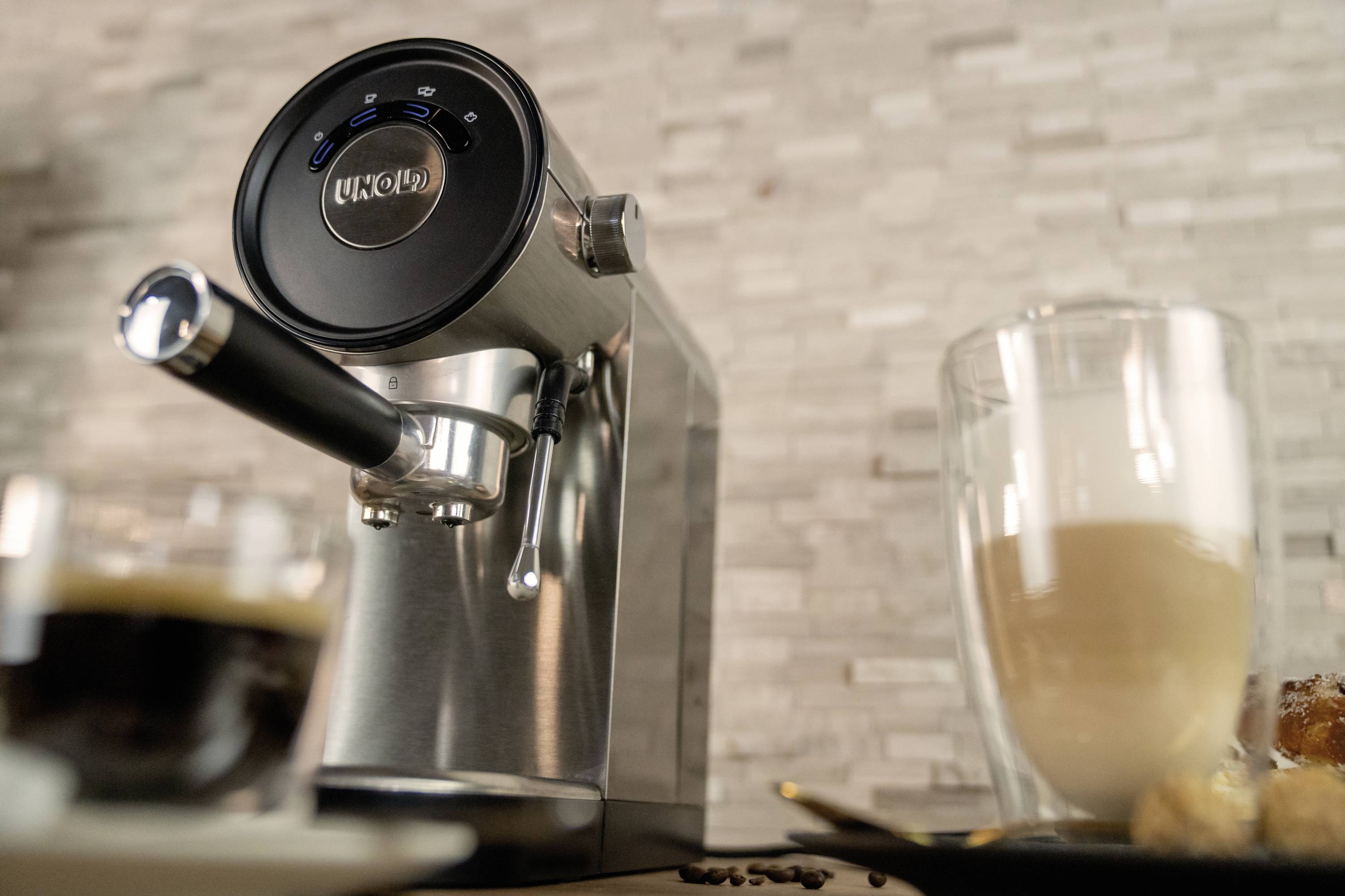 Stříbrný espresso přístroj s černým ovládacím panelem, vedle něj z poloviny naplněná sklenice latté na podnosu. Pozadí tvoří kamenné dlaždice.