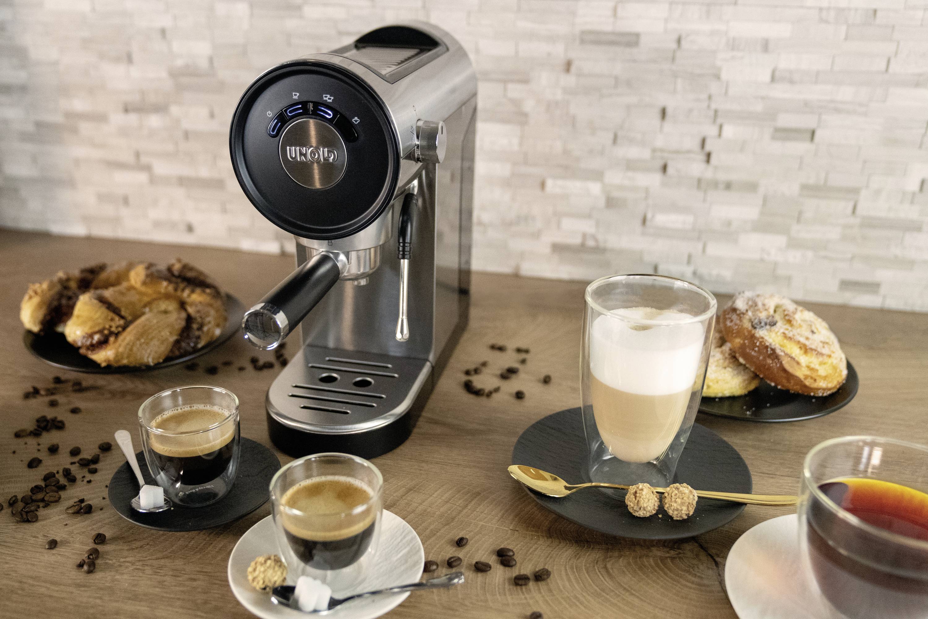 Stříbrný espresso přístroj na stole, obklopený šálky s espressem a latte macchiatem, pečivem a kávovými zrny.