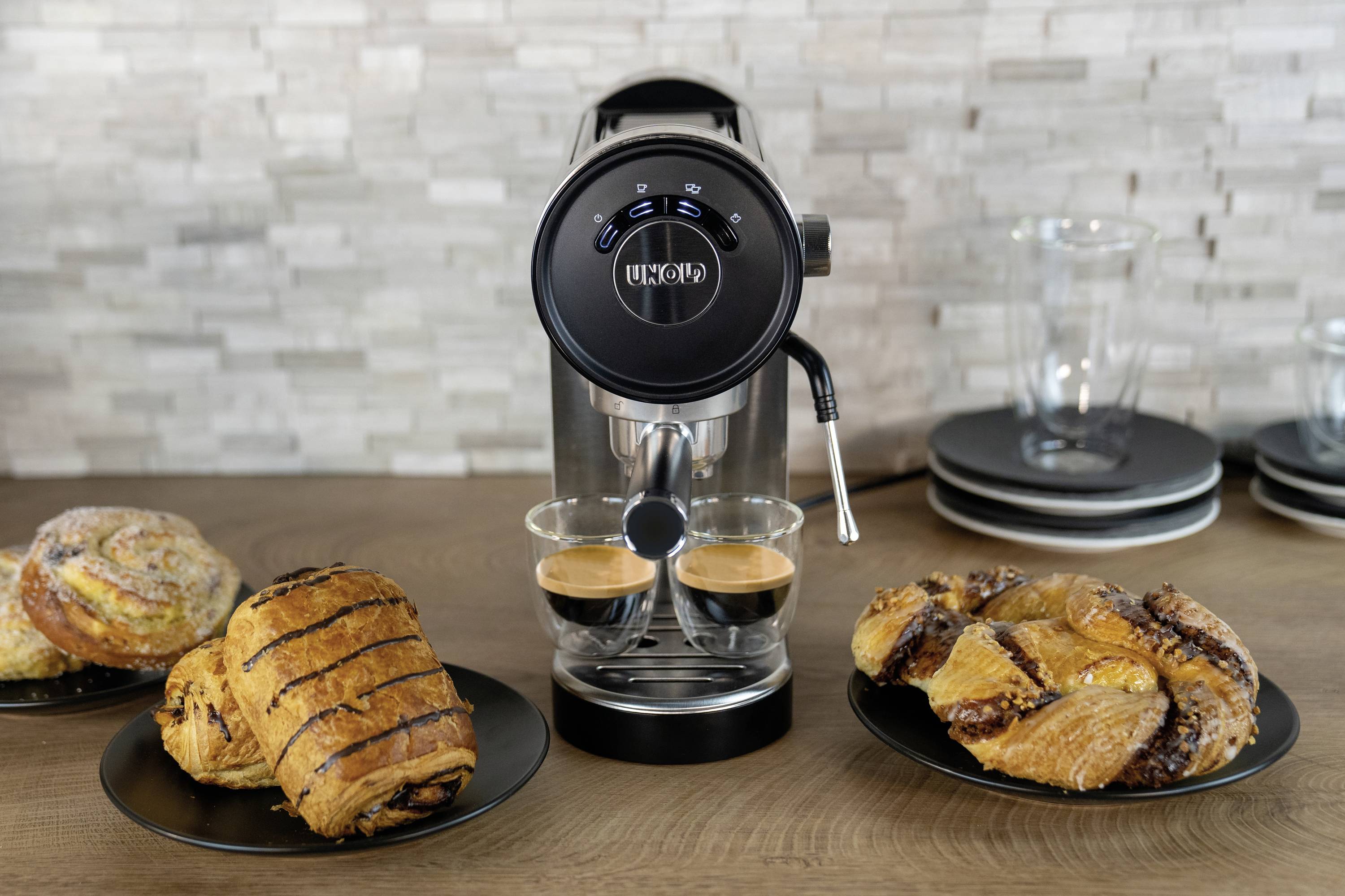 Kávový přístroj při přípravě dvou espres, lemovaný různými druhy pečiva na talířích; moderní kuchyň v pozadí.