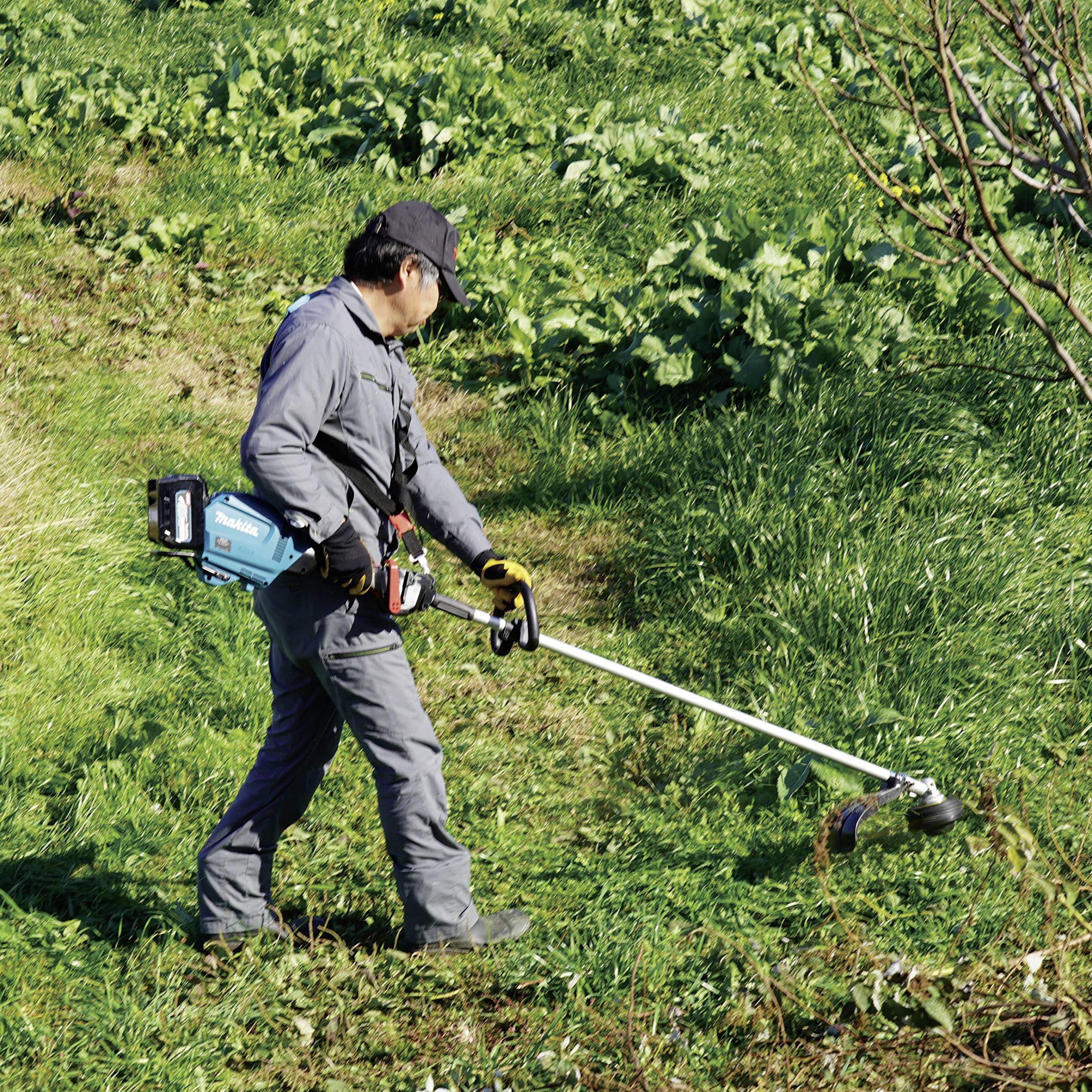 Muž v pracovním oblečení seká elektrickým křovinořezem trávu na svahu. V pozadí jsou vidět zelené rostliny.