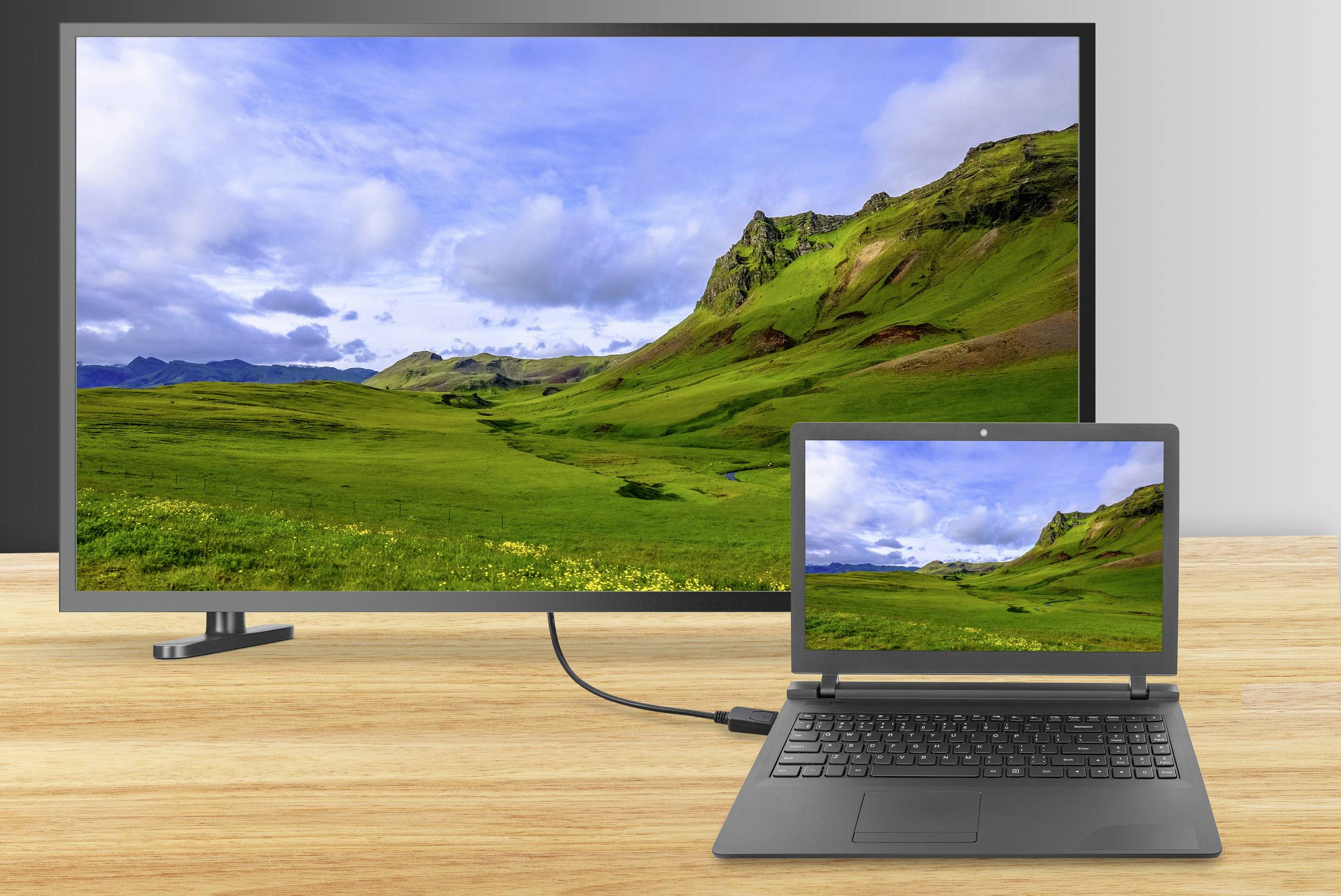 Laptop je připojen kabelem k velkému monitoru, oba zobrazují krajinu se zelenými kopci a modrou oblohou.