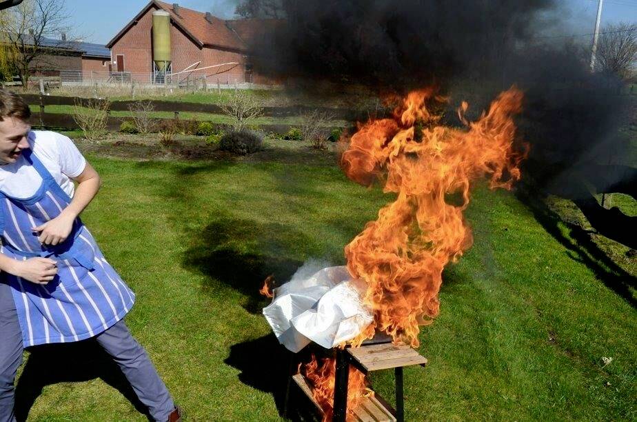Muž v pruhované zástěře stojí v zahradě a vypadá překvapeně, zatímco z grilu stoupají dramatické plameny a nad ním se valí kouř.