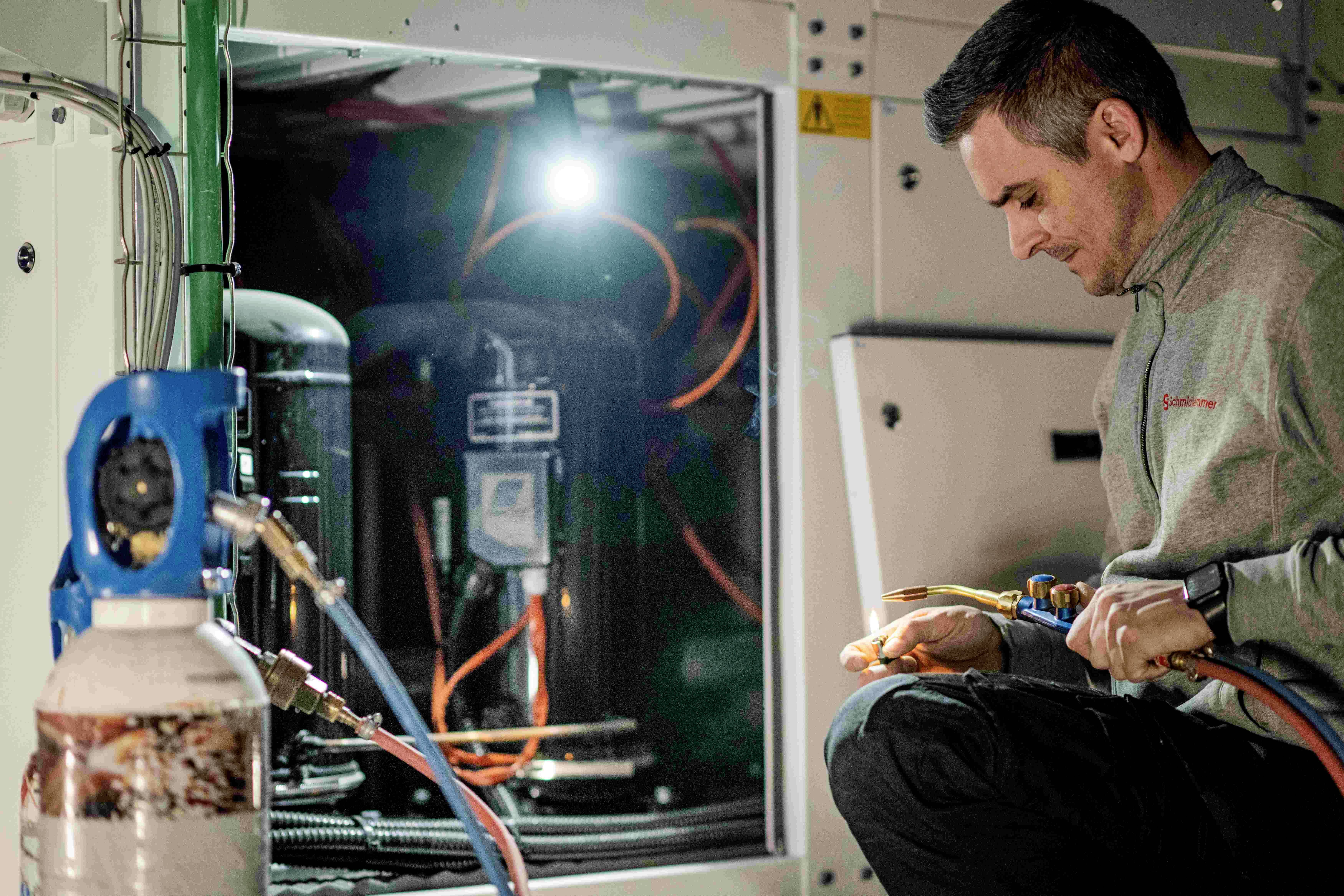 Technik se soustředěně věnuje práci s nástroji na stroji uvnitř technické místnosti, osvětlené lampou.