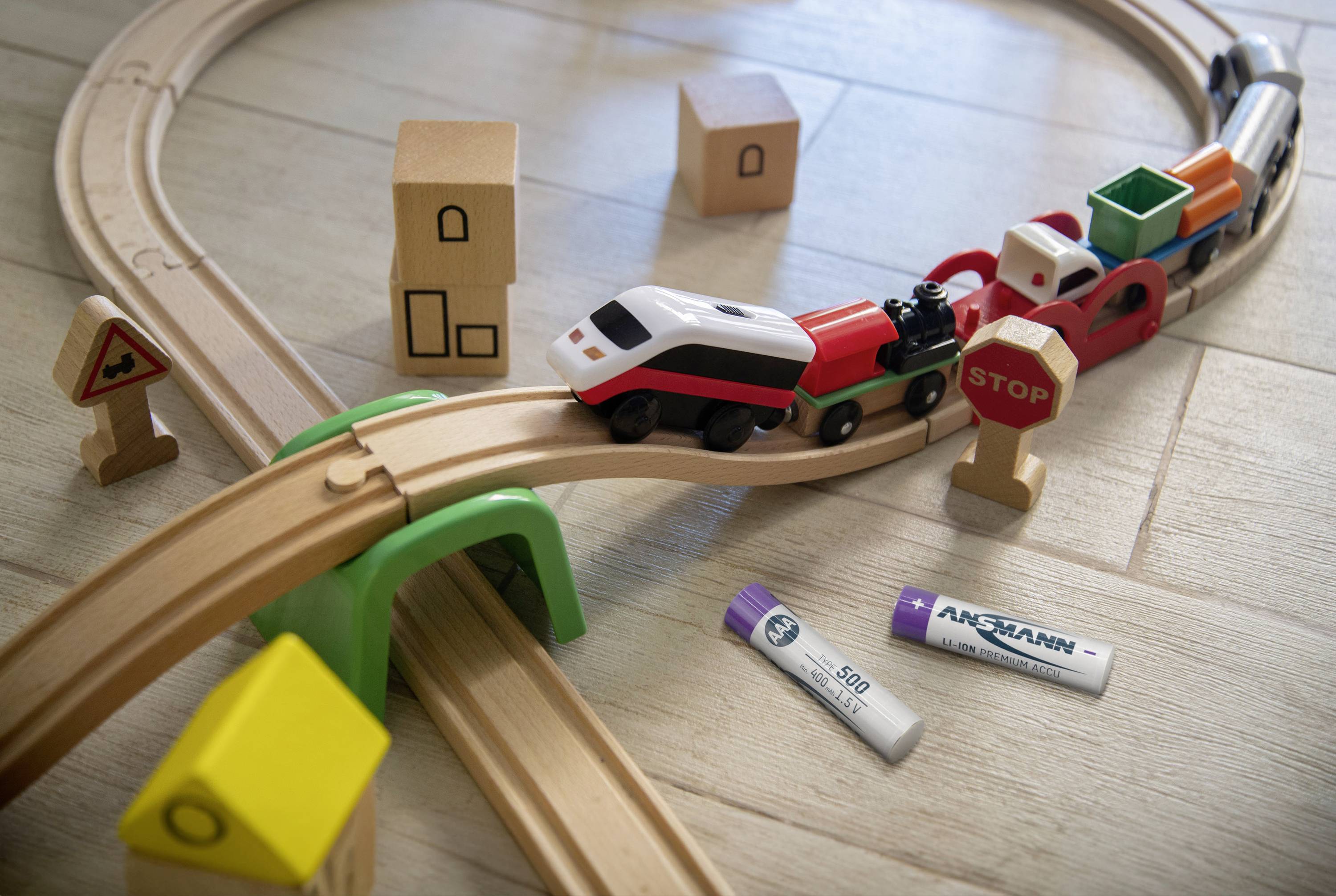 Dřevěný vláček jede po zvýšené koleji hračkářskou krajinou a míjí dopravní značky, obklopen bateriemi na světlé dřevěné podlaze.