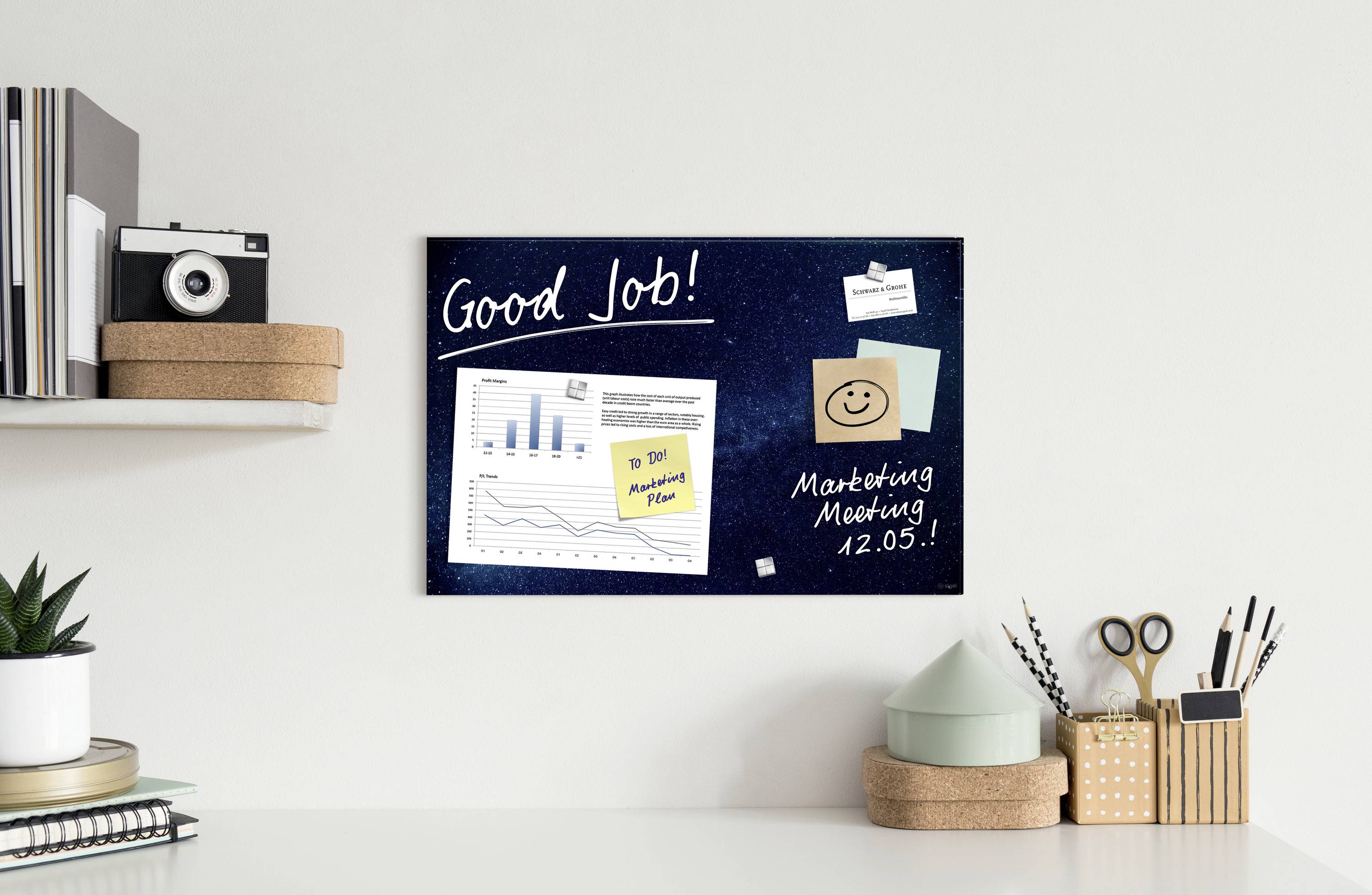Na psacím stole stojí police s fotoaparátem a rostlinami. Na stěně visí nástěnka s poznámkami, grafiky a texty 'Good Job!' a 'Marketing Meeting 12.05.'.