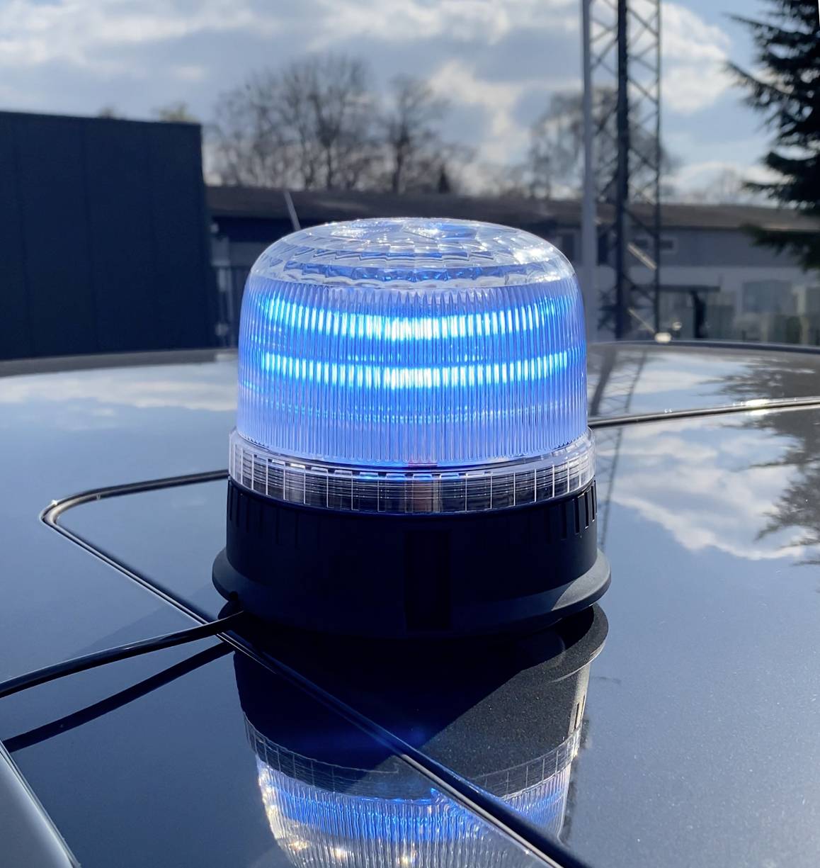 Modrý maják na střeše automobilu odrážející sluneční světlo. V rozmazaném pozadí je vidět budova a strom.