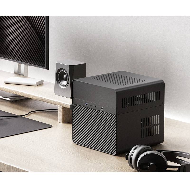 Na psacím stole stojí moderní černý mini počítač vedle sluchátek a reproduktorů. Vedle je viditelný monitor.