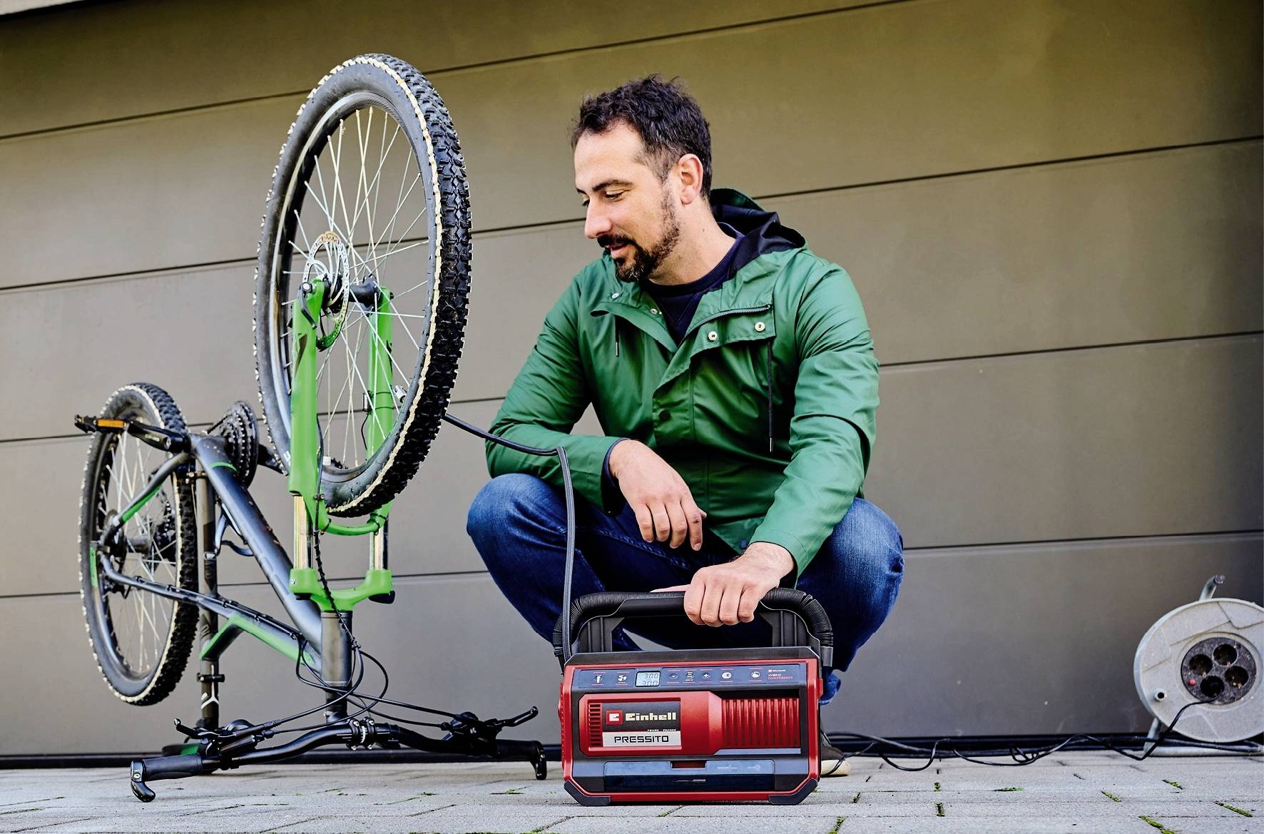 Muž v zelenémanoráku opravuje převrácené jízdní kolo pomocí červeného elektrického nástroje.