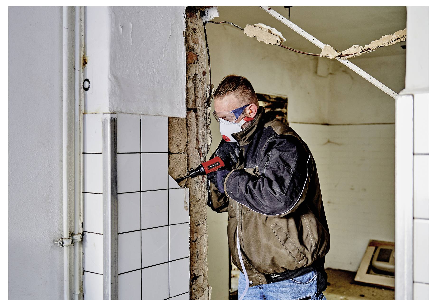 Pracovník v ochranné masce a brýlích vrtá do obkládané stěny během demolice nebo renovace v částečně vyutaženém interiéru.