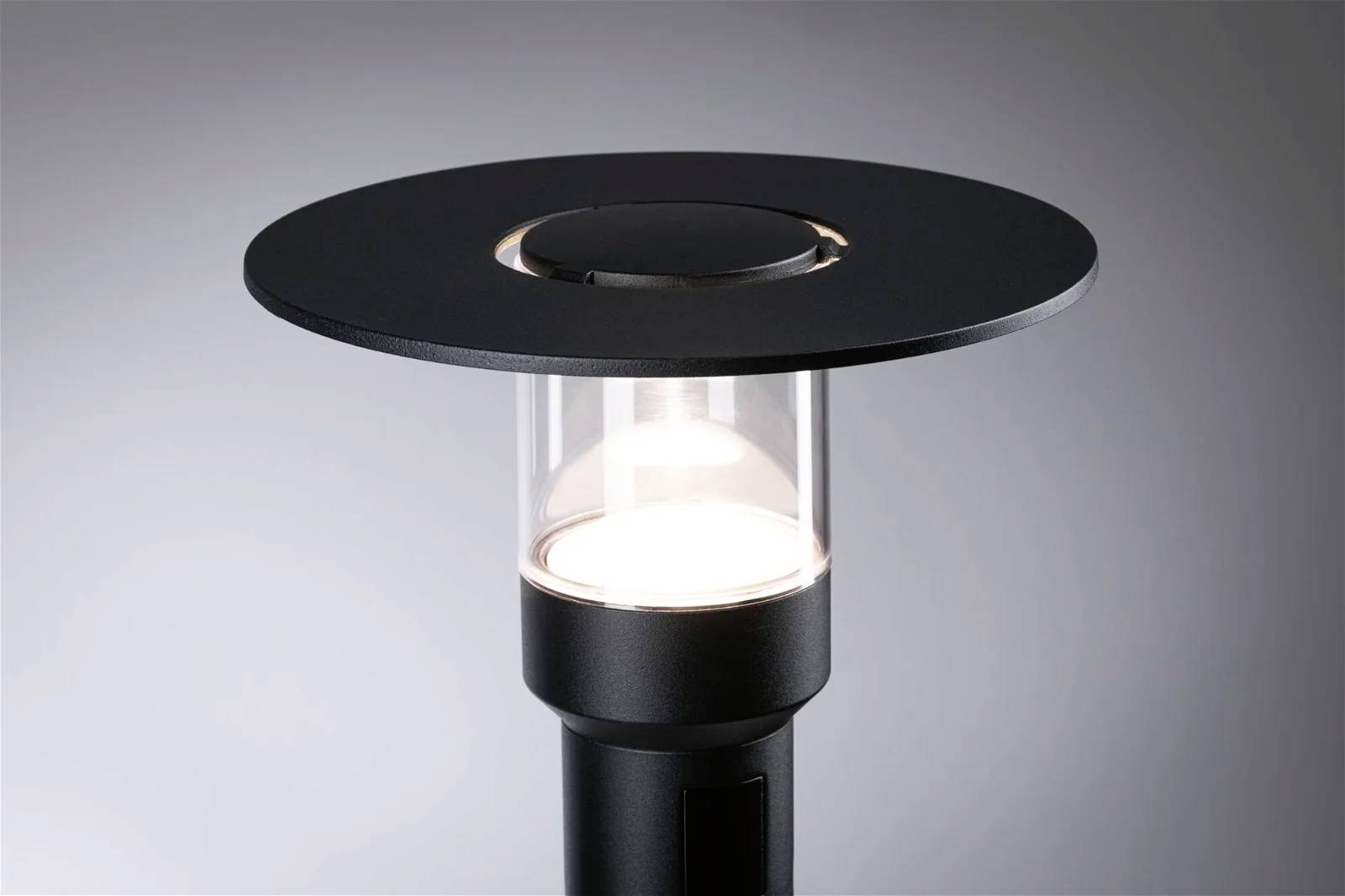 Černá moderní stojací lampa s průhledným skleněným válcem a kulatým, plochým stínidlem, osvětlená v minimalistickém prostoru.
