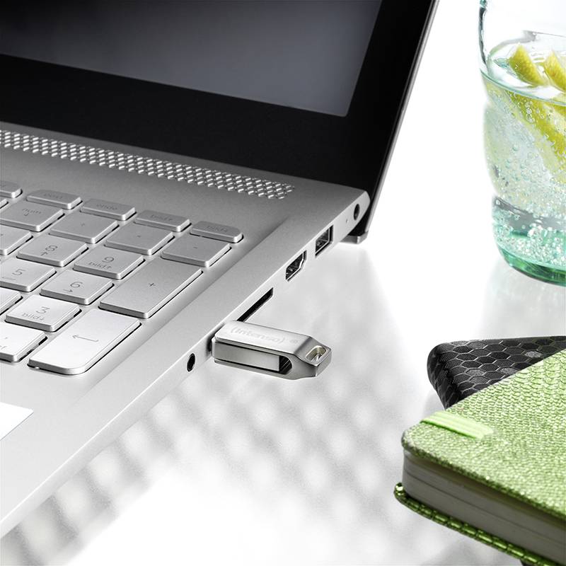 Notebook s připojeným USB flash diskem leží na stole. Vedle něj je poznámkový sešit a sklenice vody s citronem.