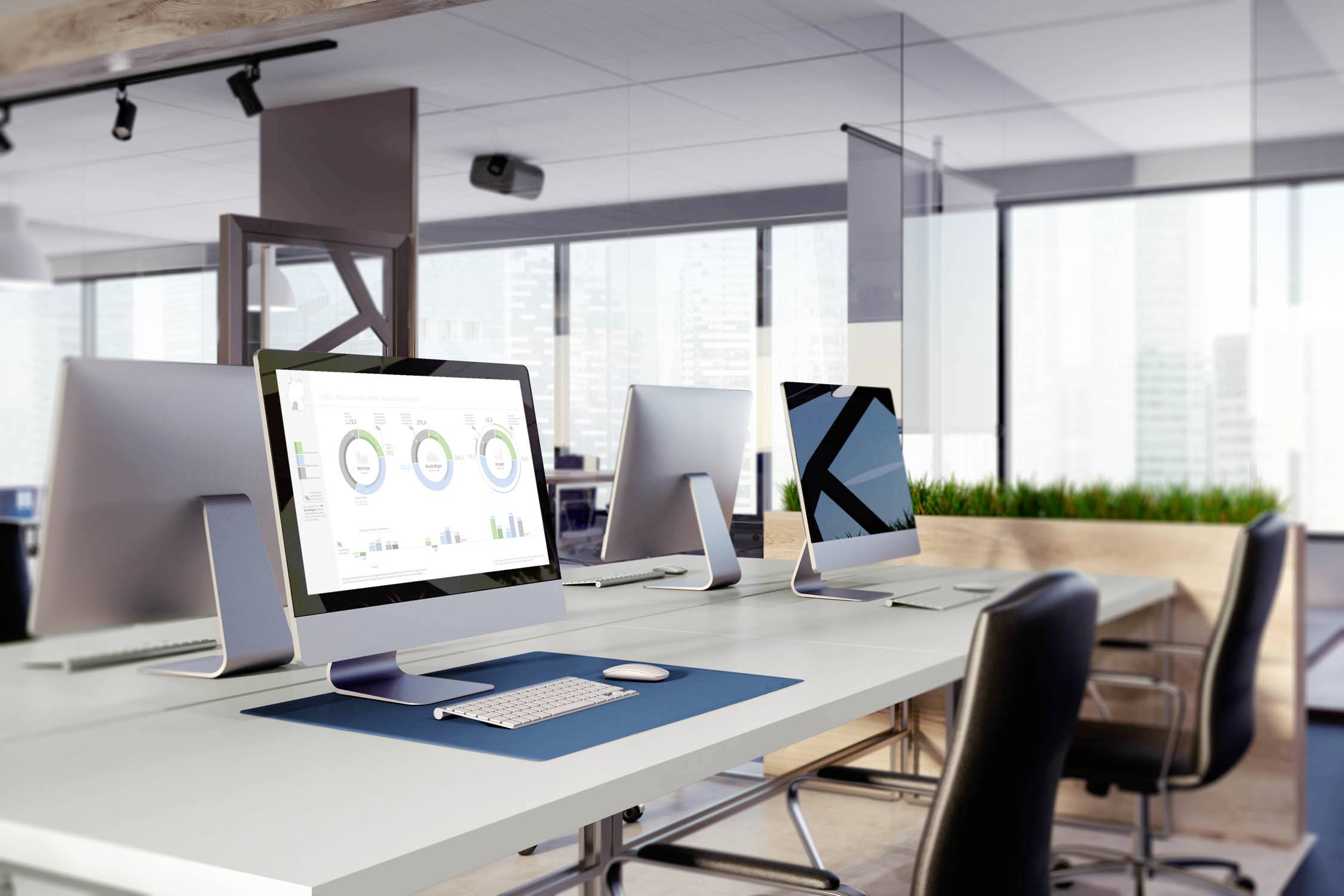 Moderní kancelářské prostory s několika počítači na pracovních stolech. Na jedné obrazovce jsou viditelné diagramy. Velká okna v pozadí.