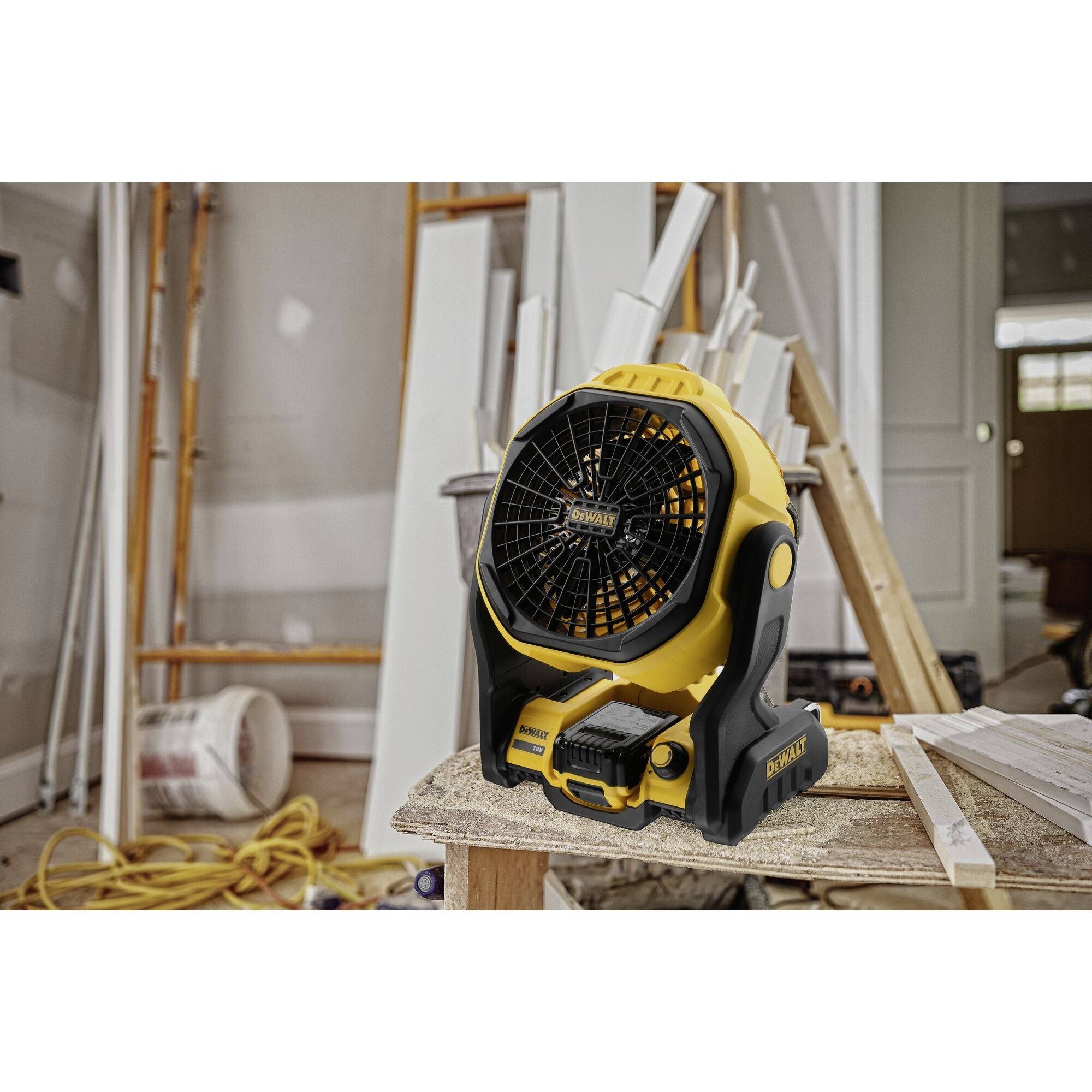 Žlutý ventilátor stojí na stole v dílně. V pozadí jsou vidět stavební materiály a lešení.