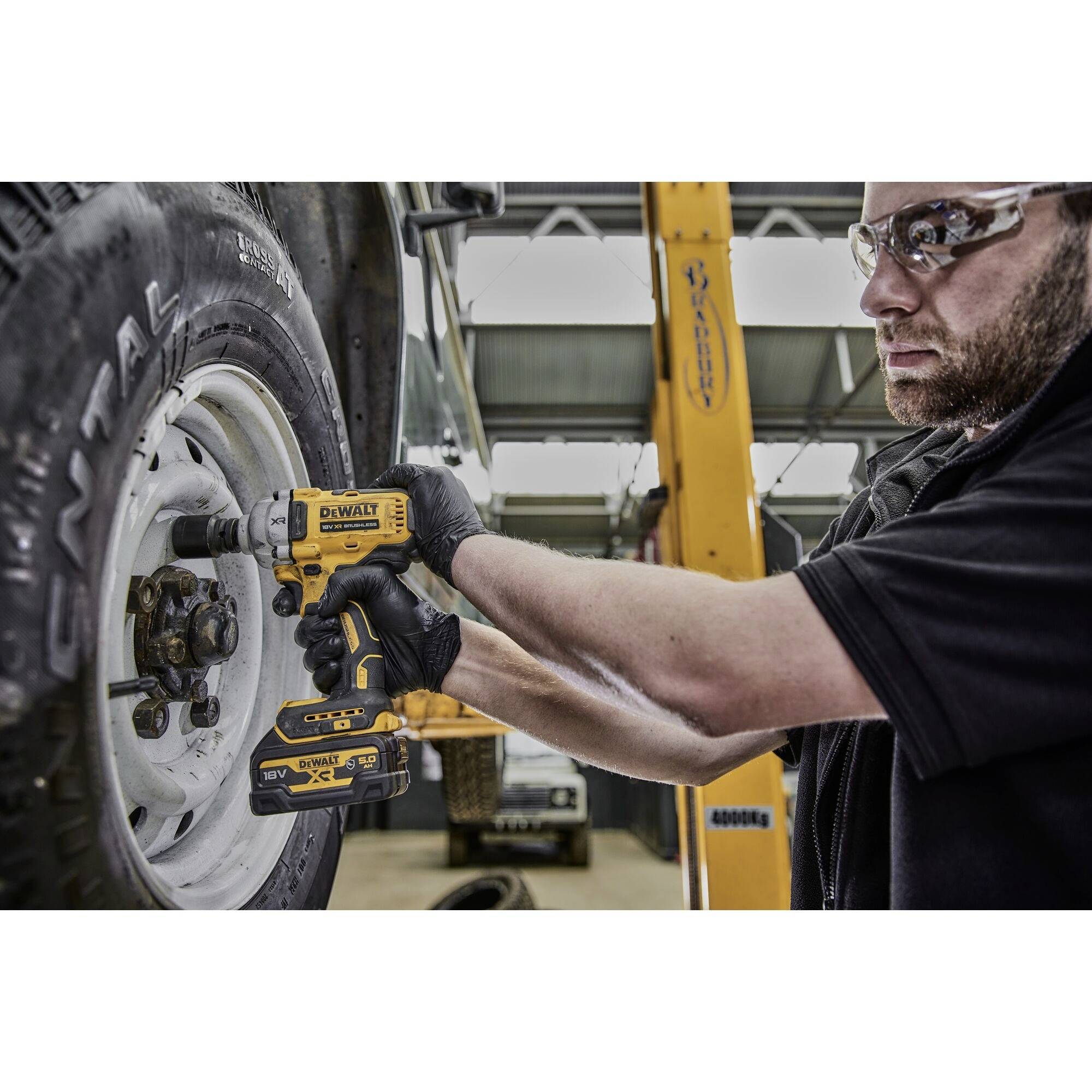 Osoba používá žlutý akumulátorový šroubovák k utažení matice kola automobilu v autoservisu. V pozadí jsou viditelné automobilové díly.