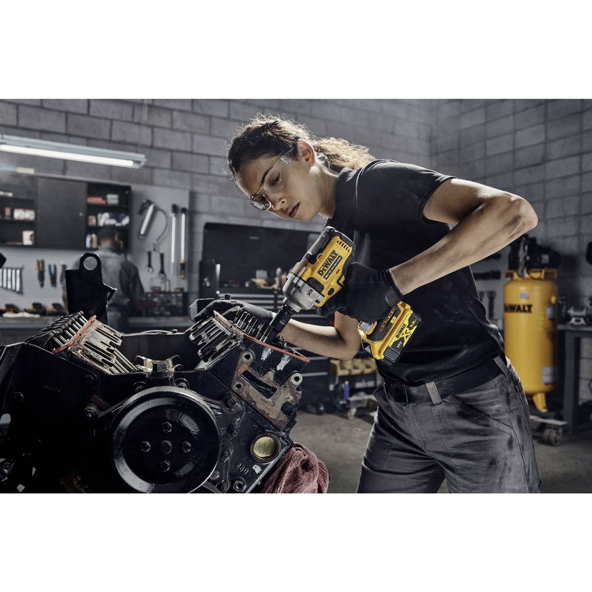 Mechanička pracuje s vrtačkou na bloku motoru v dílně. V pozadí jsou viditelné nástroje a vybavení.