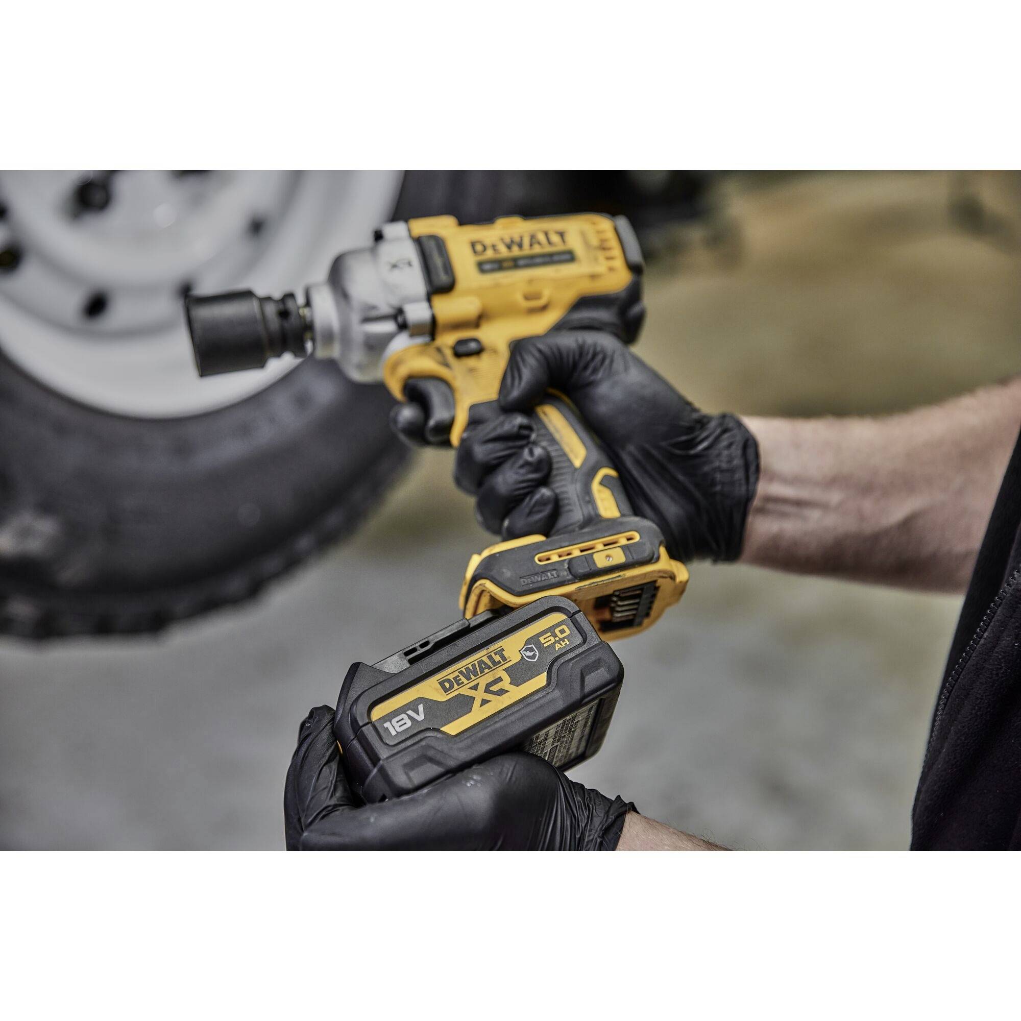 Akumulátorový šroubovák značky DEWALT s vloženou baterií, držený osobou v černých rukavicích. V pozadí je viditelný automobilový pneumatika.