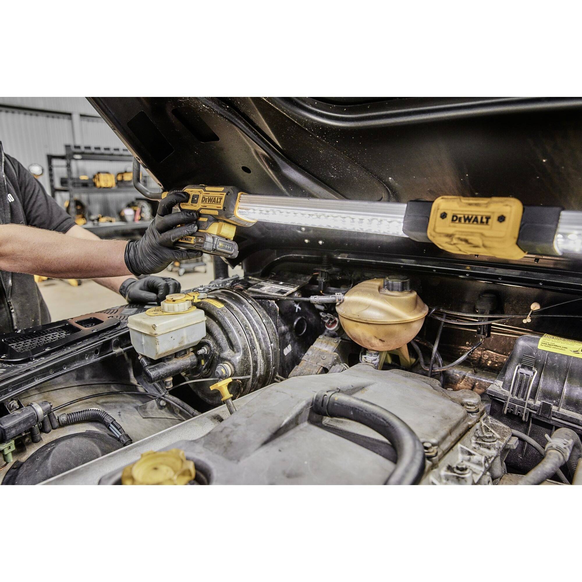 Mechanik používá lampu DeWalt k osvětlení motoru vozidla. Kapota je otevřená a jsou viditelné nástroje.