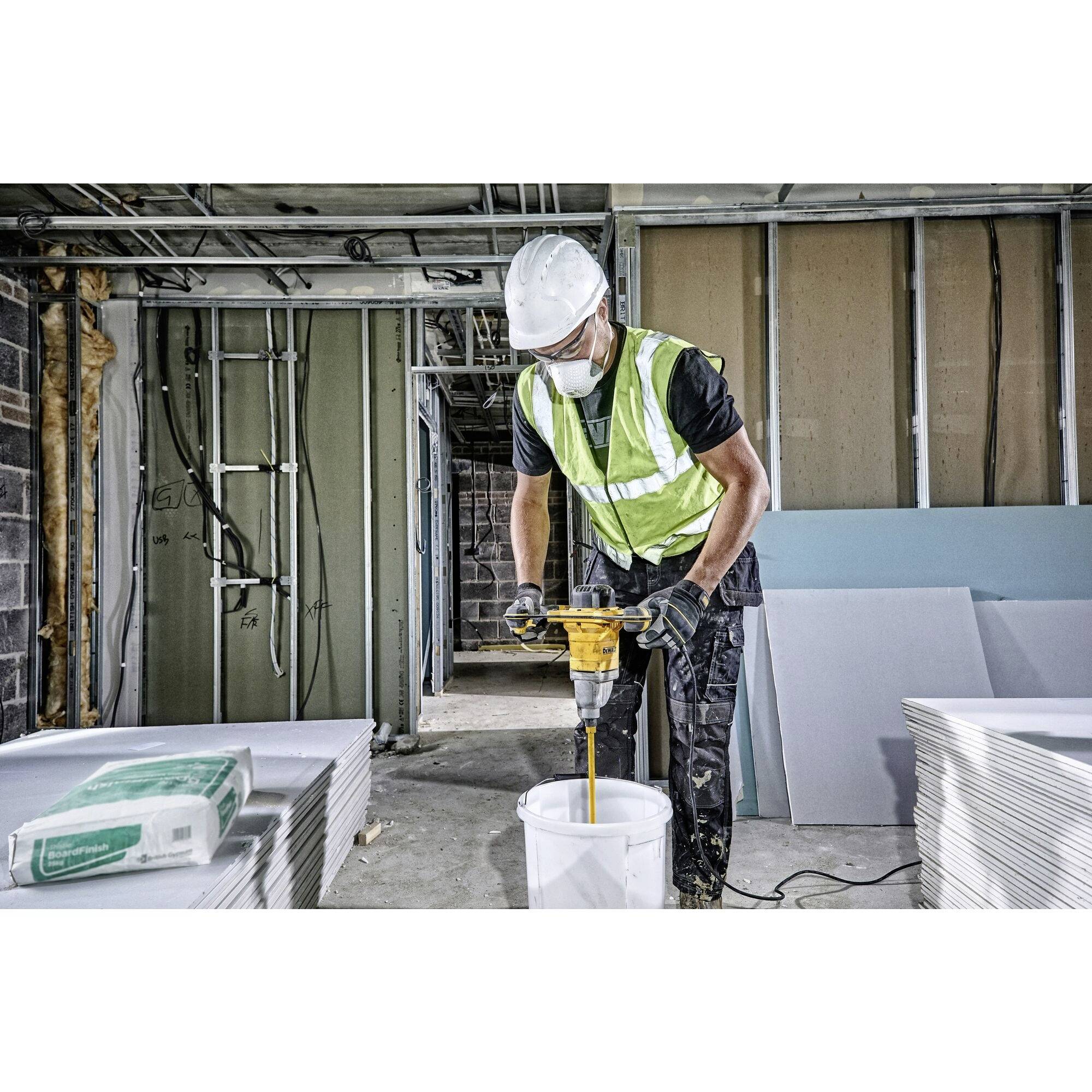 Dělník v helmě a vestě míchá cement ve vědru v nedokončené místnosti plné stavebních materiálů.