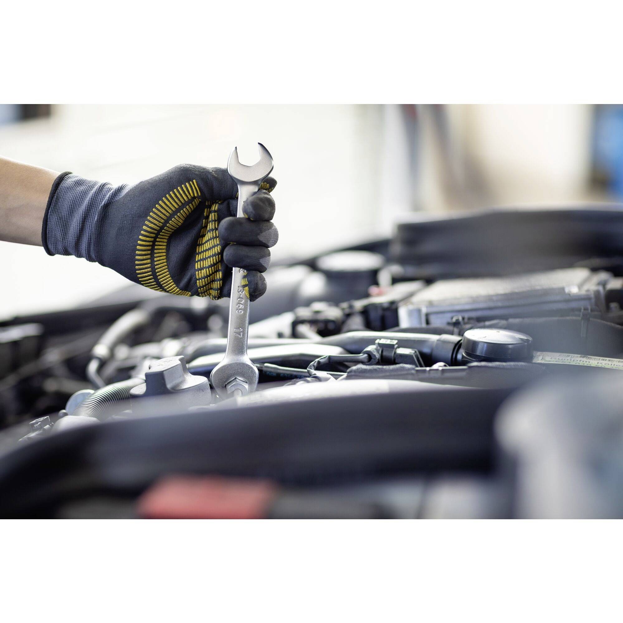 Mechanik v rukavici drží klíč nad otevřeným motorovým prostorem, zatímco pracuje na automobilu.