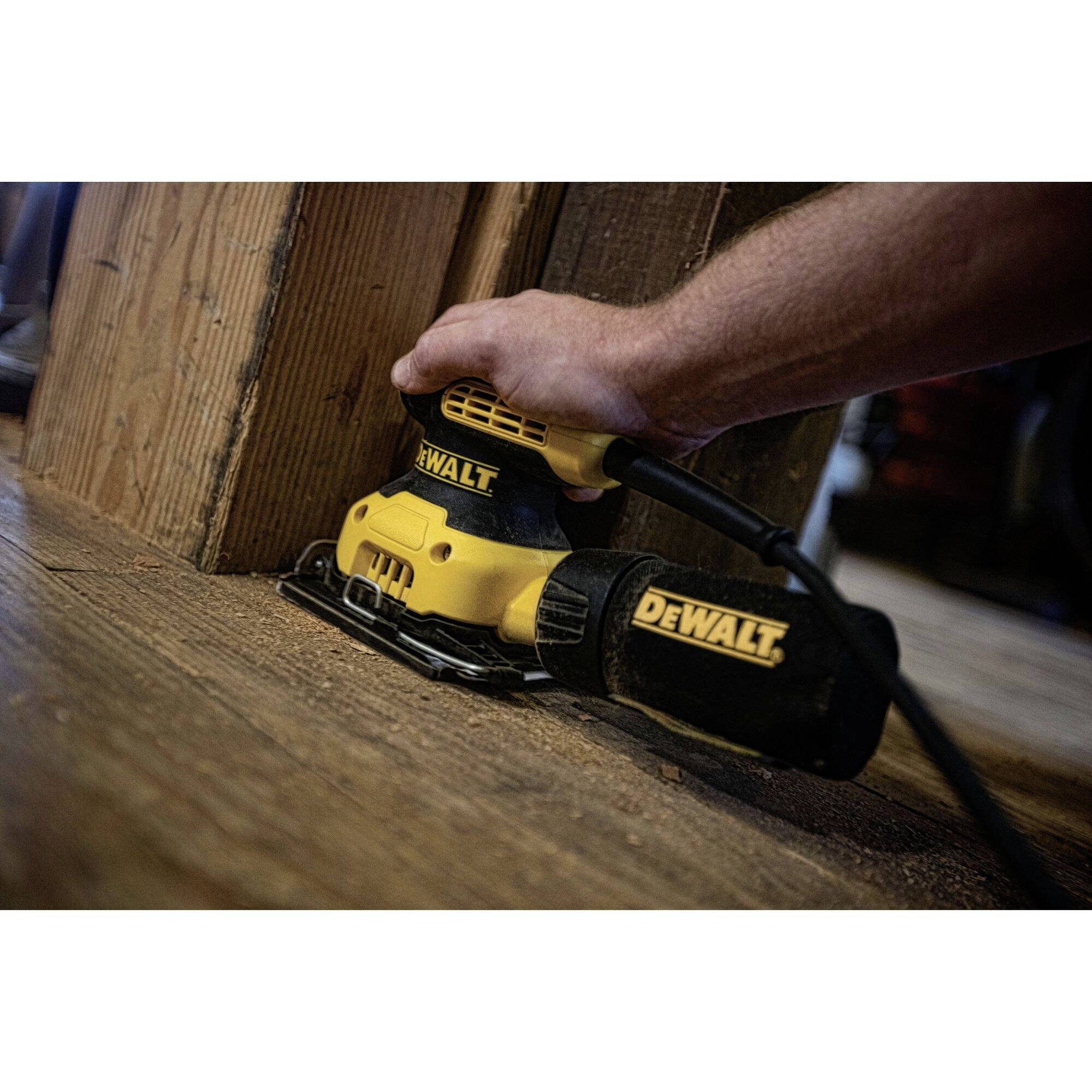 Jedna ruka drží žlutou elektrickou brusku značky DeWalt, která brousí dřevěný díl v místnosti.