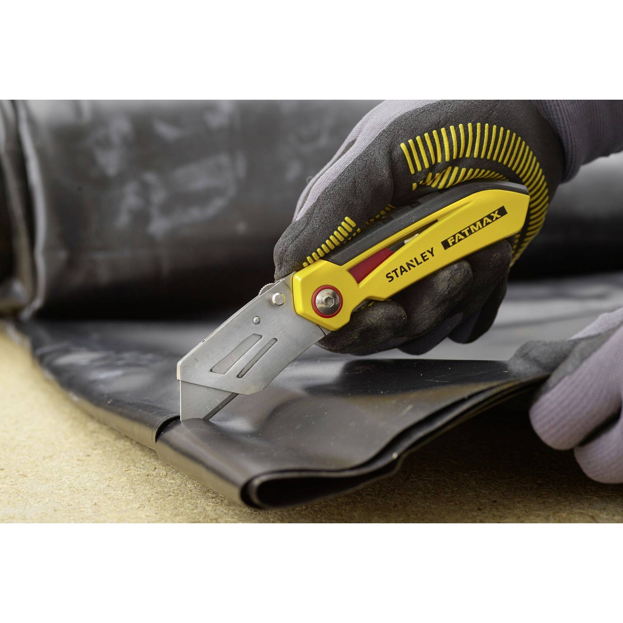 Ruka v černé rukavici řeže tmavý materiál na stole pomocí nože Stanley FatMax.
