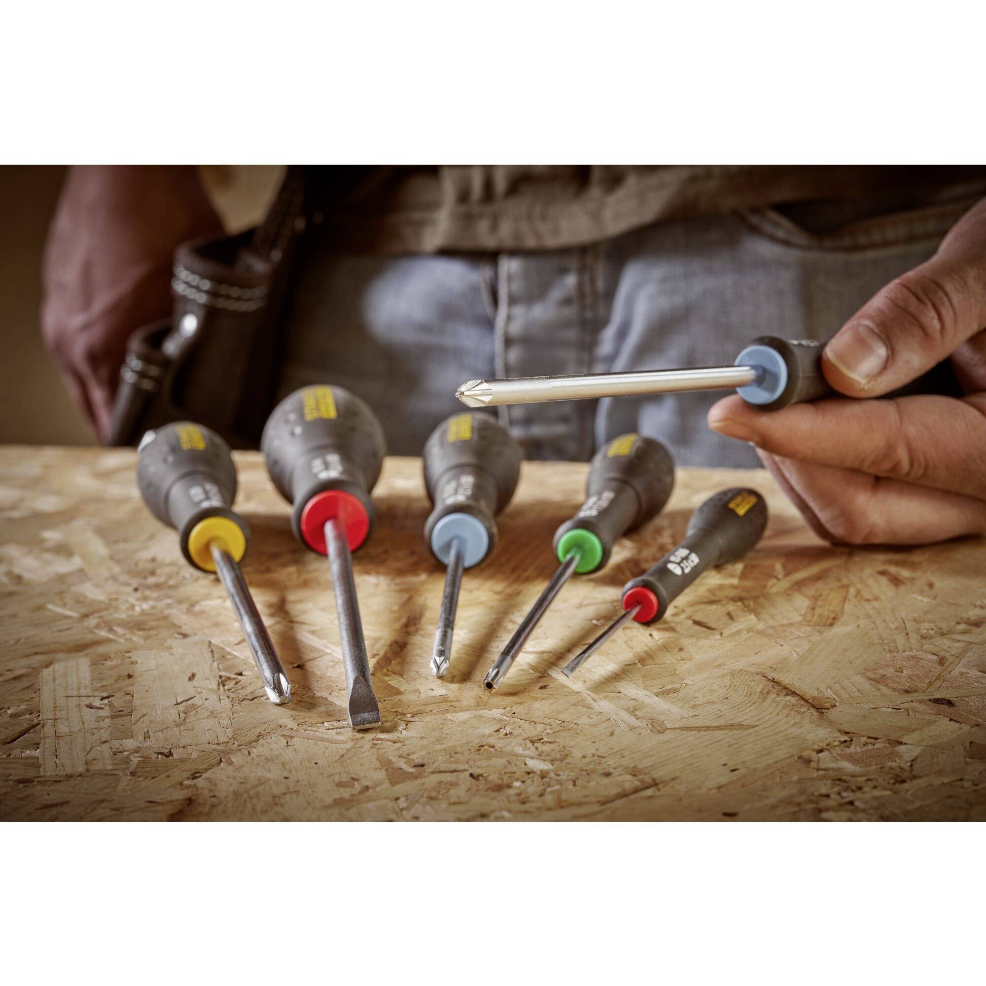 Sada pěti šroubováků v různých barvách na dřevěném povrchu. Jedna ruka drží šroubovák.