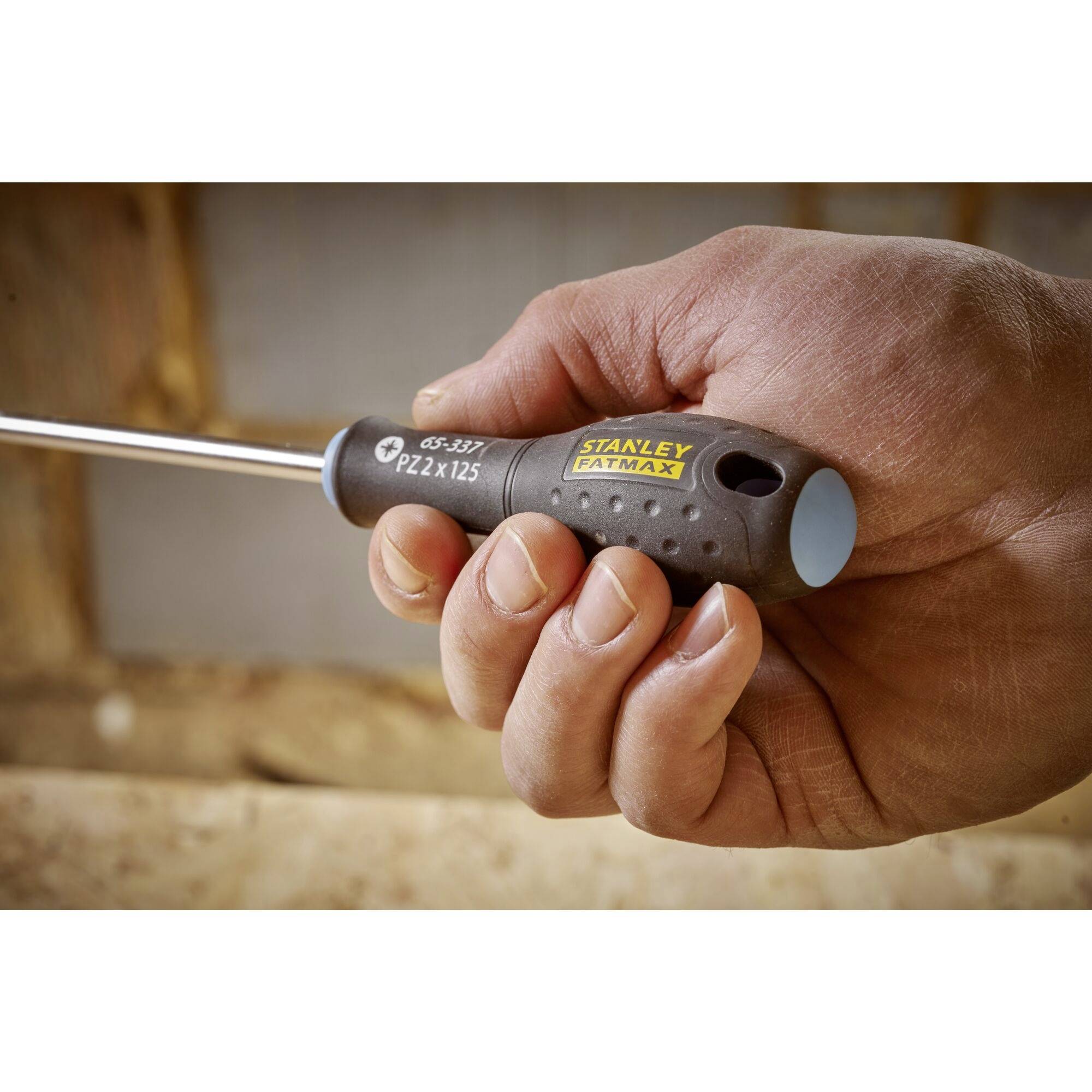 Jedna ruka drží šroubovák značky 'Stanley Fatmax', připravený k použití. Hrot je namířen doleva.