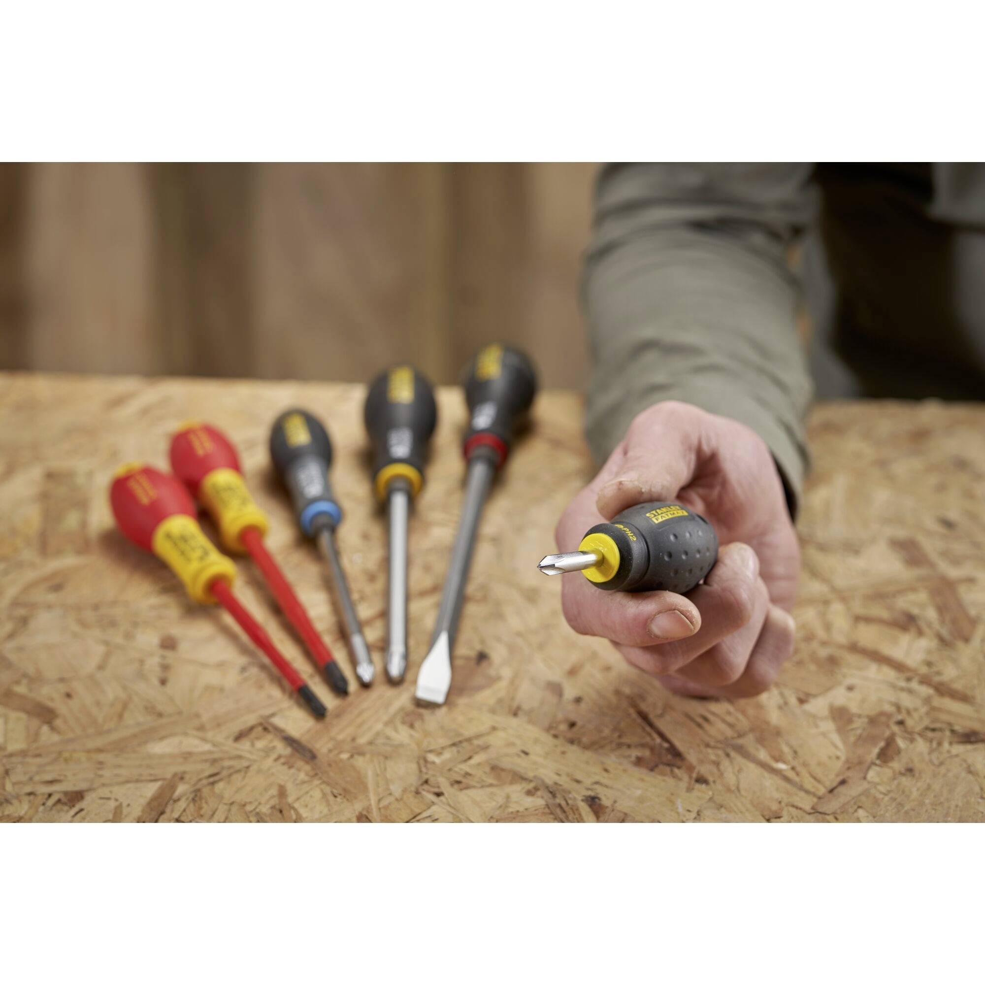 Ruka drží malý šroubovák, zatímco v pozadí leží několik dalších šroubováků na dřevěném stole.