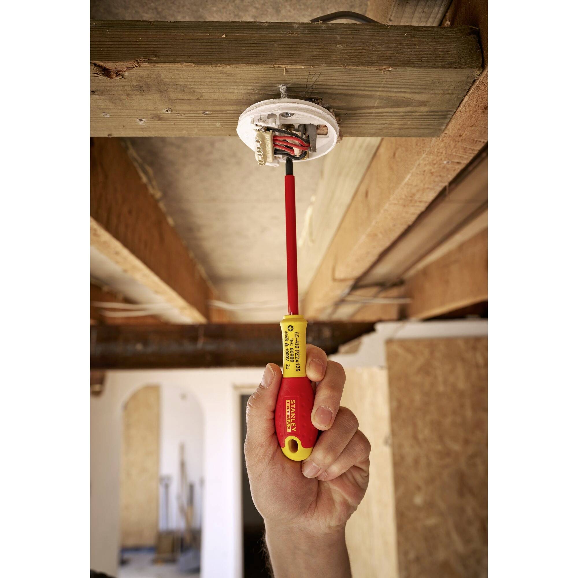 Jedna ruka upevňuje žlutý šroubovák stropní svítidlo na dřevěném trámovém stropě v nedokončené místnosti.