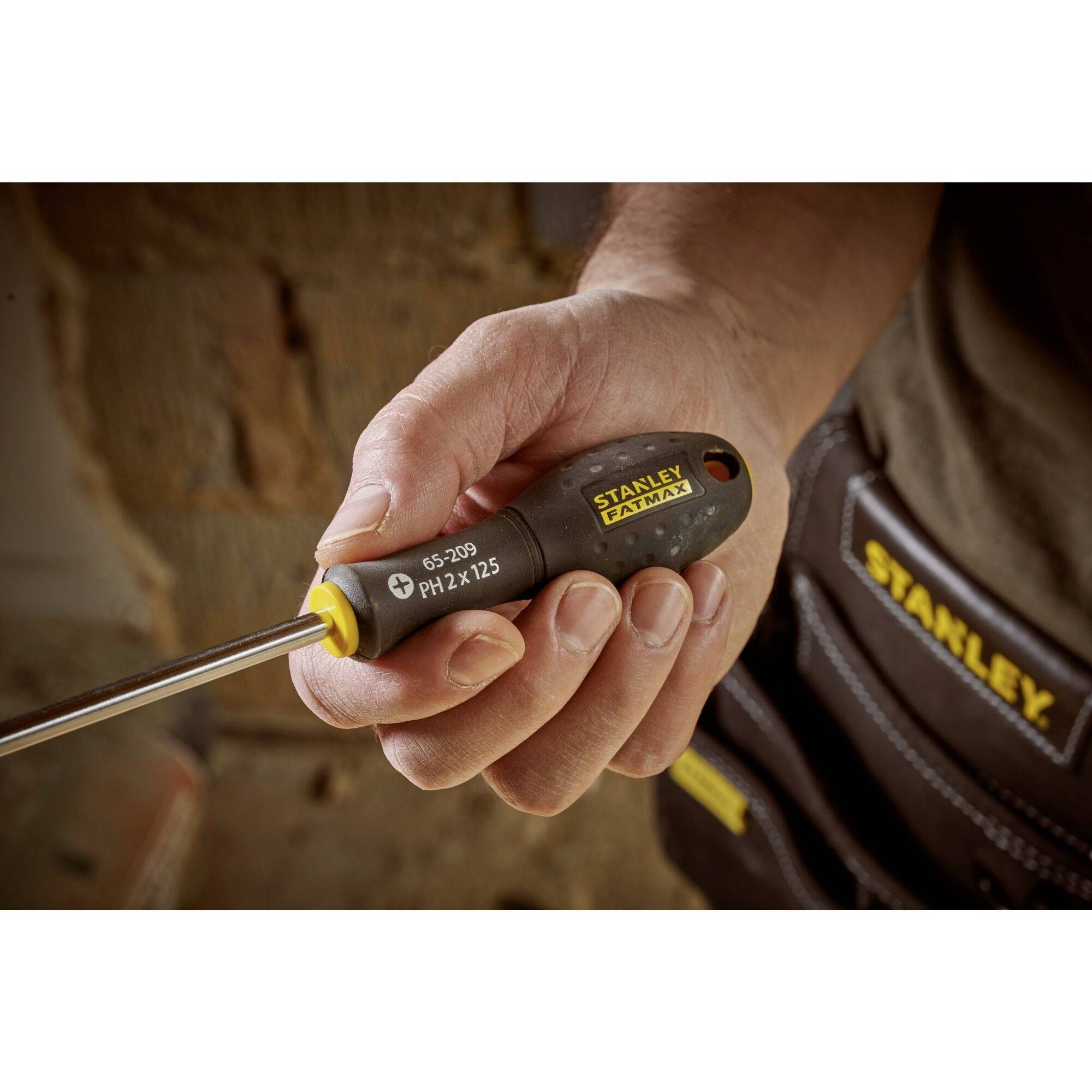 Osoba drží šroubovák s černou rukojetí. V pozadí je viditelná brašna na nářadí.