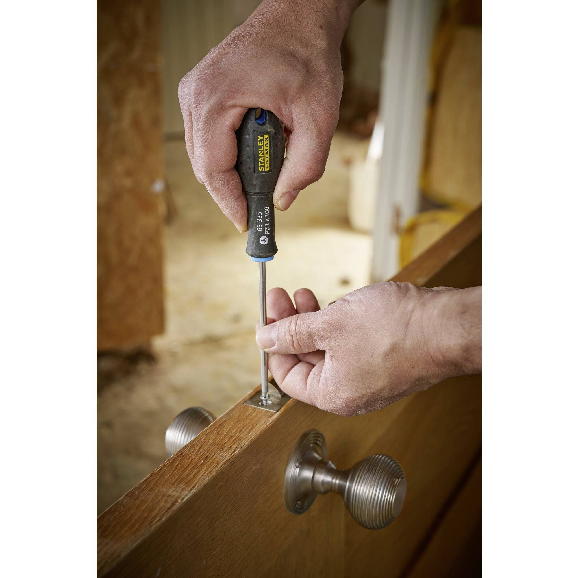 Člověk utahuje dveřní závěs šroubovákem. Dřevěný povrch a kovová klika jsou viditelné.