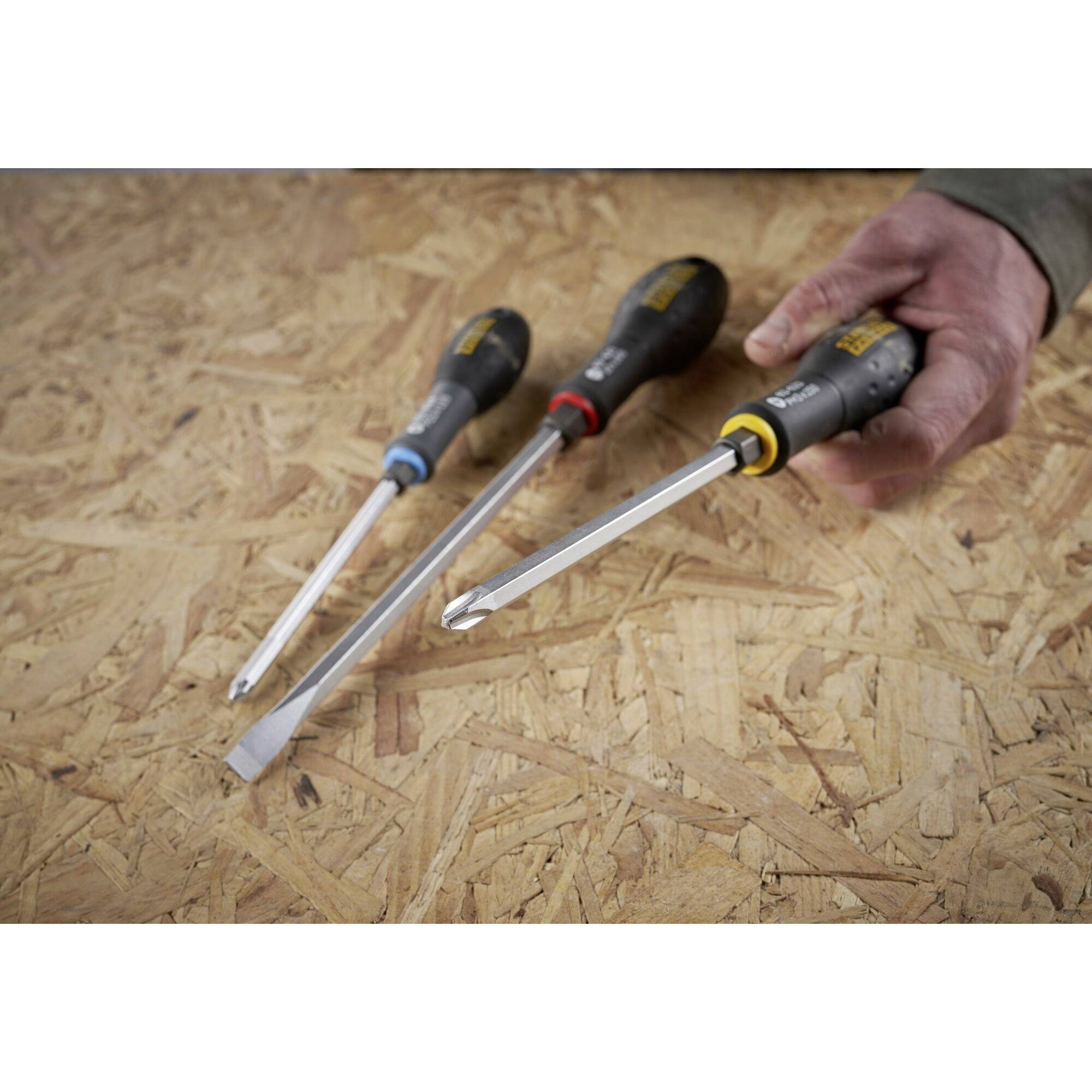 Tři šroubováky na dřevěné desce, jeden šroubovák je držen rukou. Různé velikosti a typy, příslušenství pro řemeslníky.