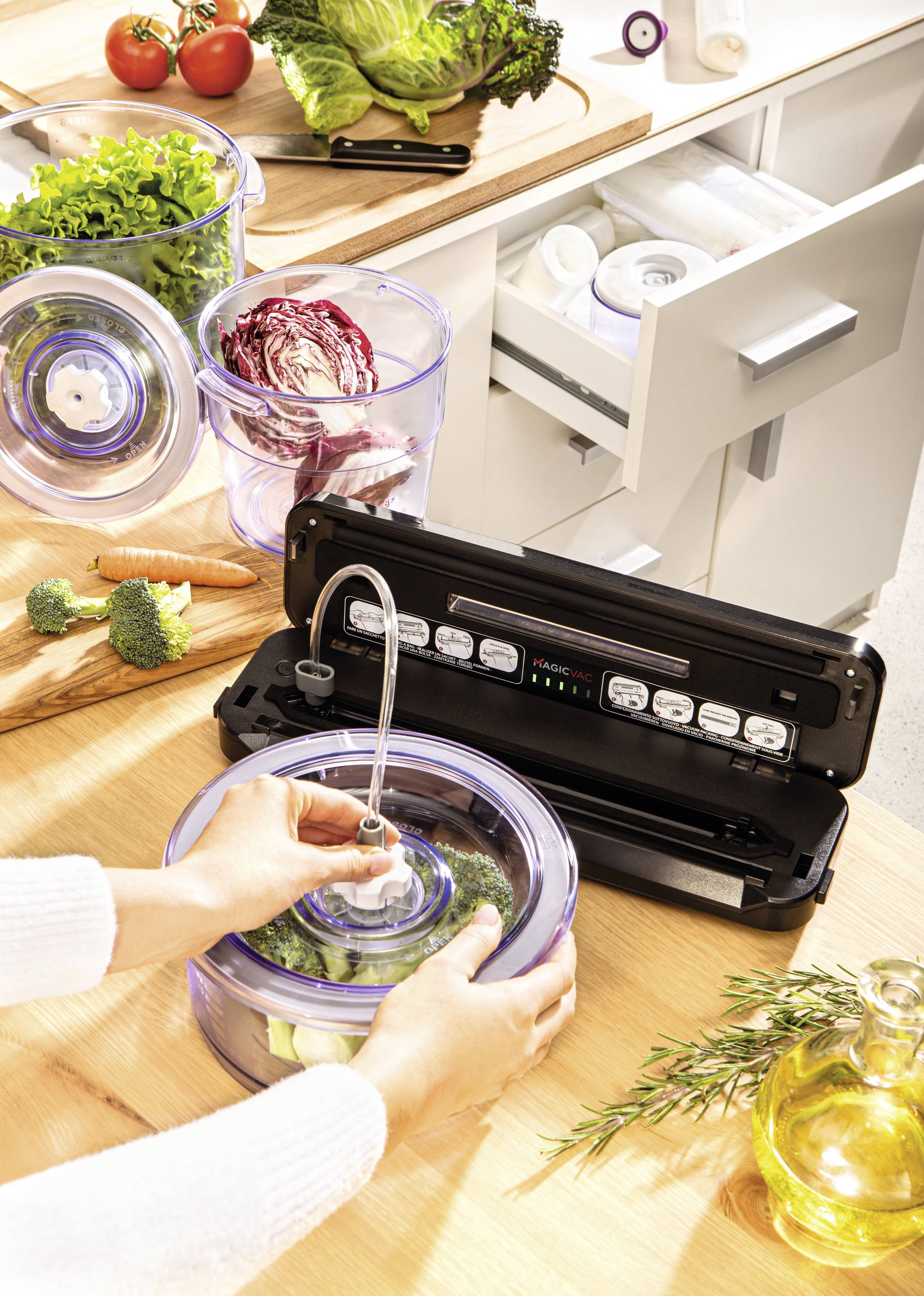 Osoba vakuuje zeleninu v nádobě pomocí vakuovačky. Na pracovní ploše leží čerstvá zelenina a kuchyňské pomůcky.